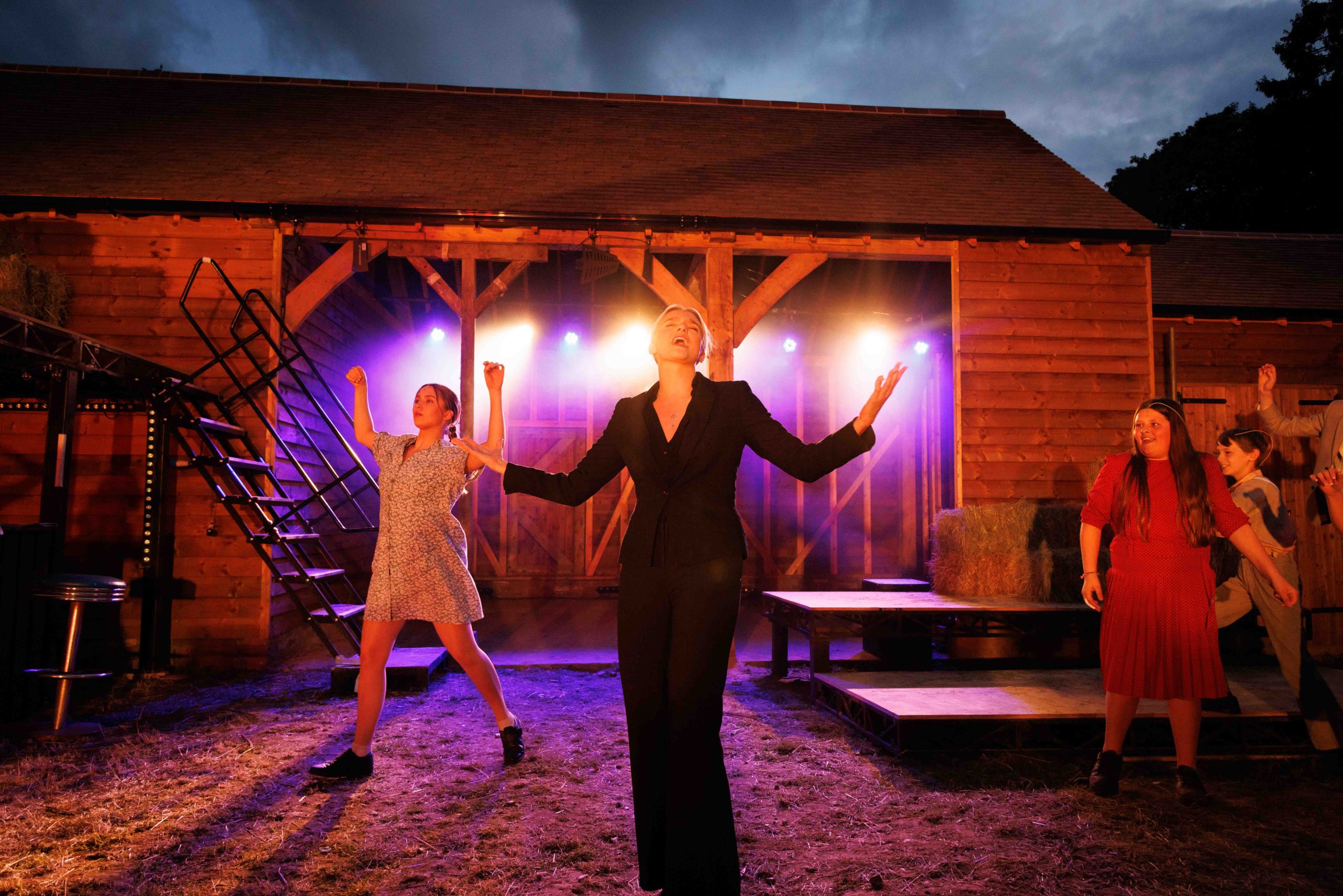 A performer in a black suit sings centre stage with arms outstretched as ensemble members dance in front of a lit wooden barn at dusk.