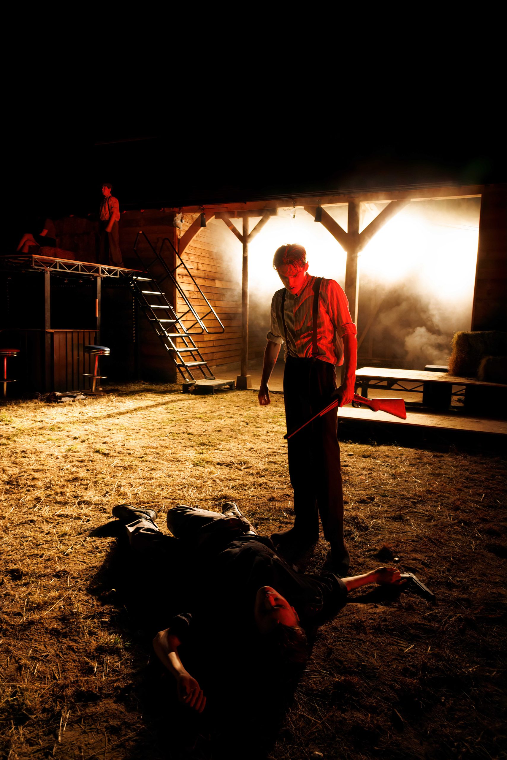 A performer stands over a figure lying on the ground in front of the lit barn set at night.