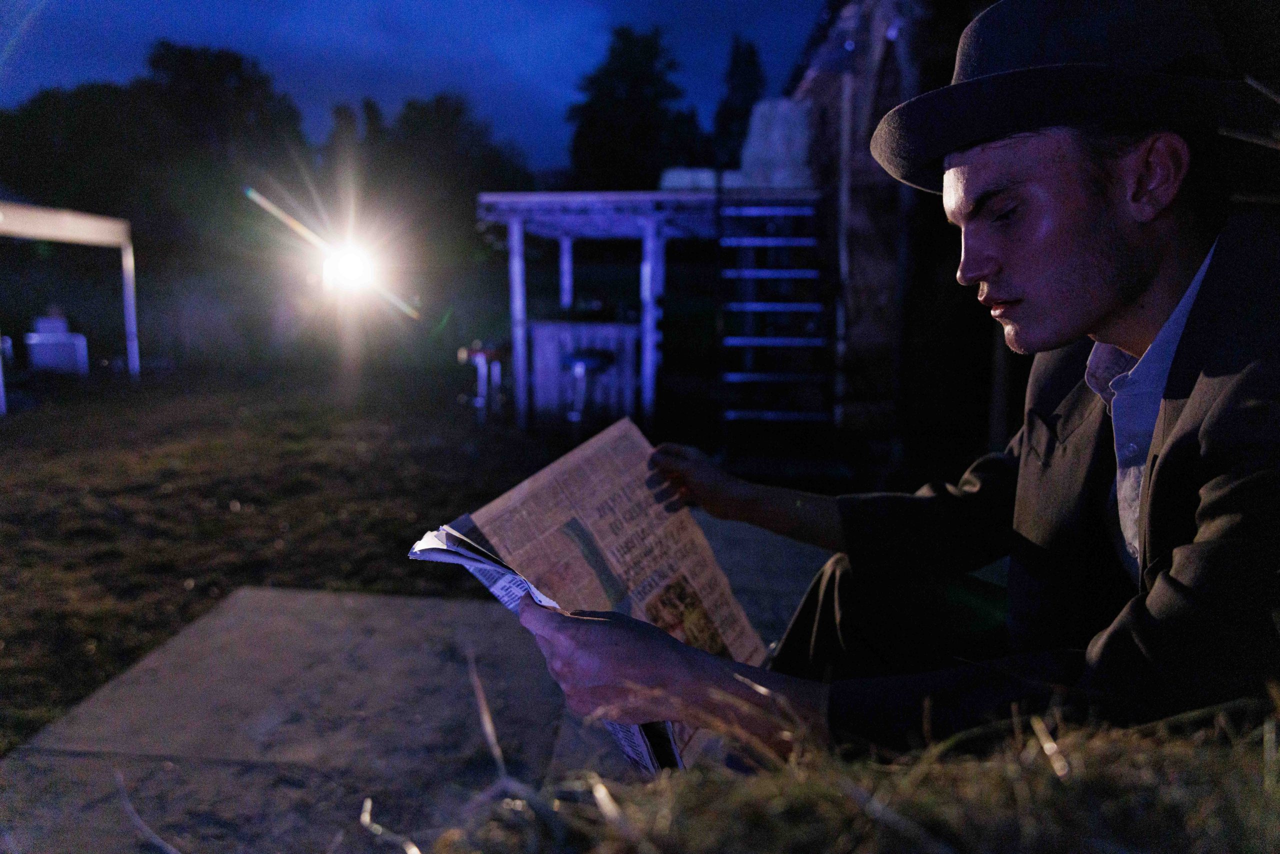 A performer in a trilby hat reads a newspaper at night on an outdoor set, with a bright light shining through the haze behind.
