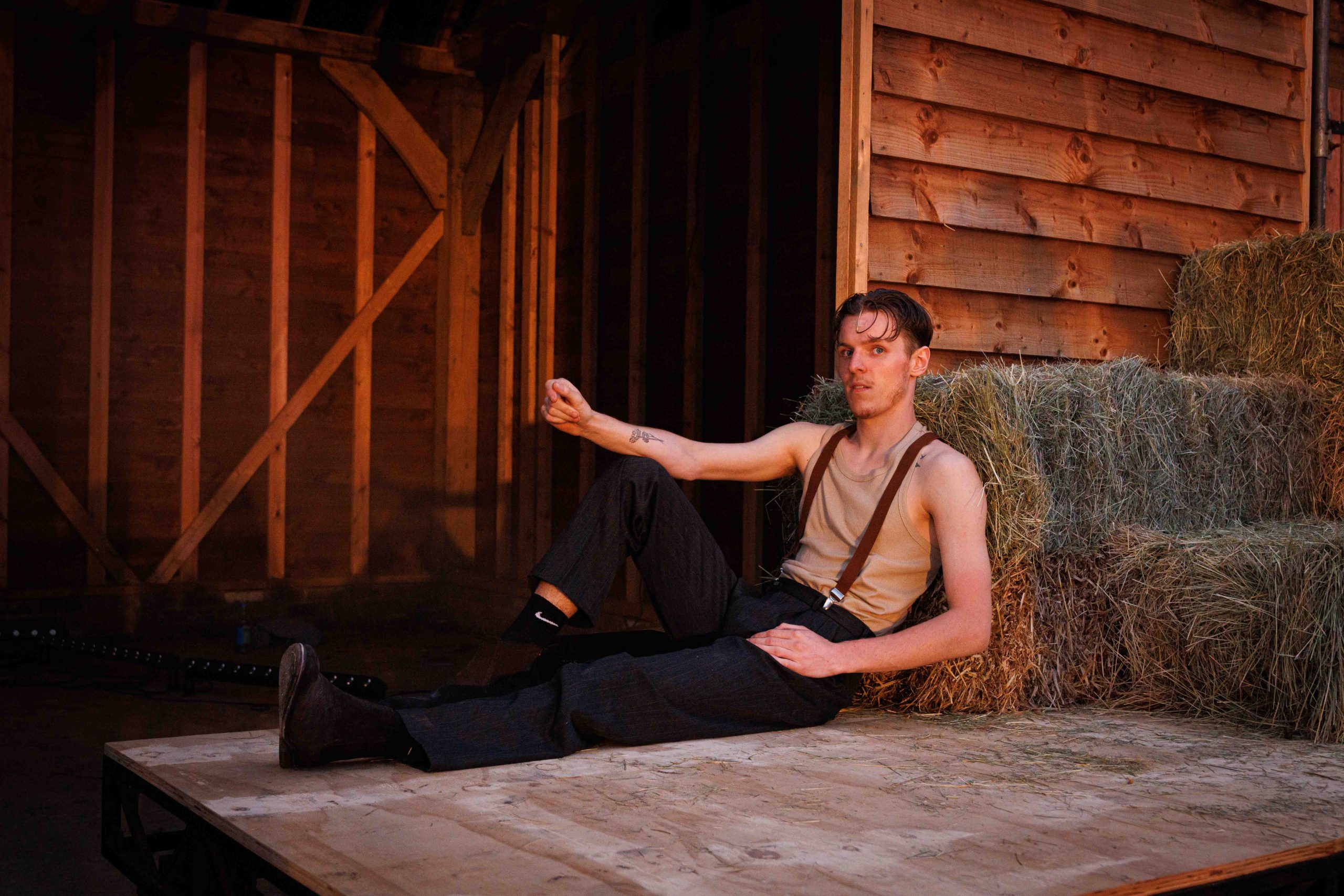 A performer in a vest and braces reclines against hay bales on a wooden barn set, lit in warm amber.