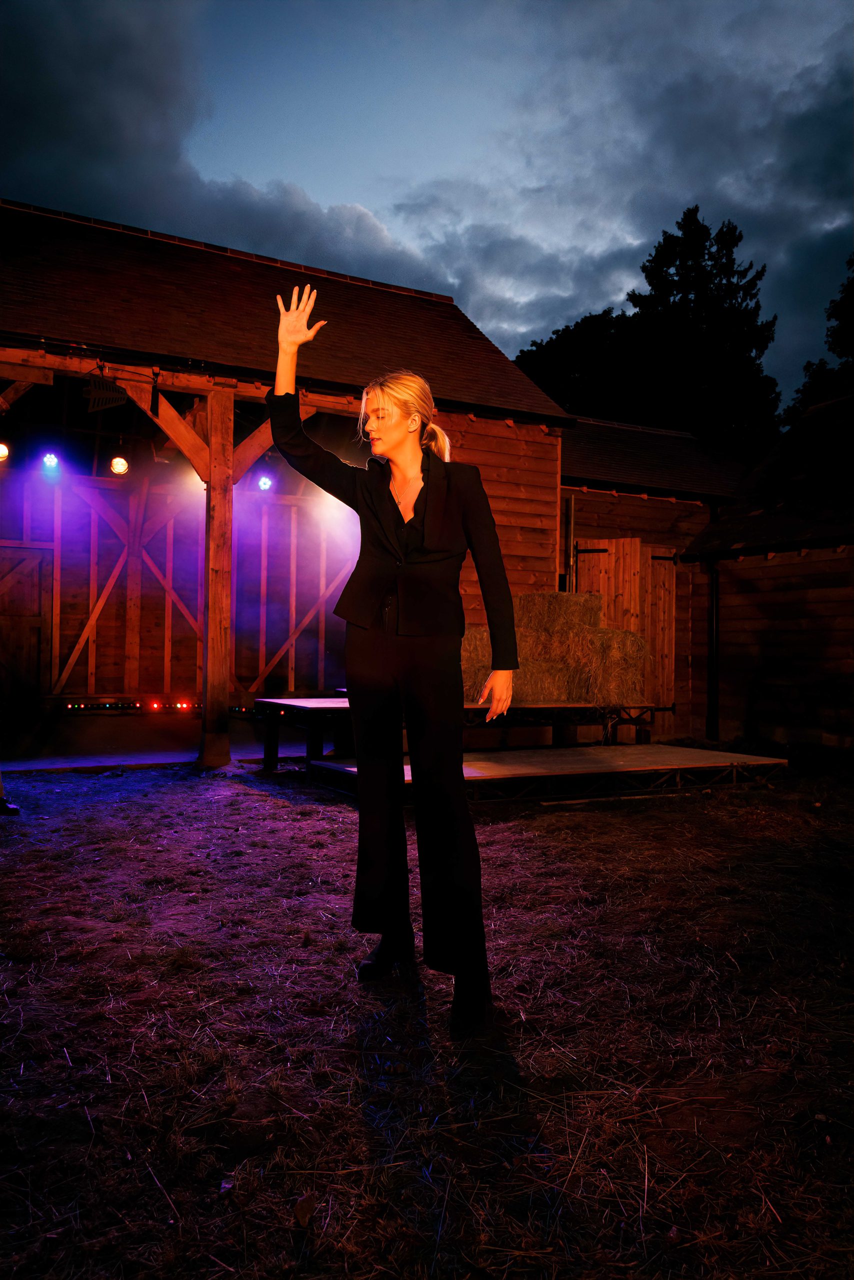A performer in a dark suit raises one hand while singing in front of the barn set under a dusk sky.
