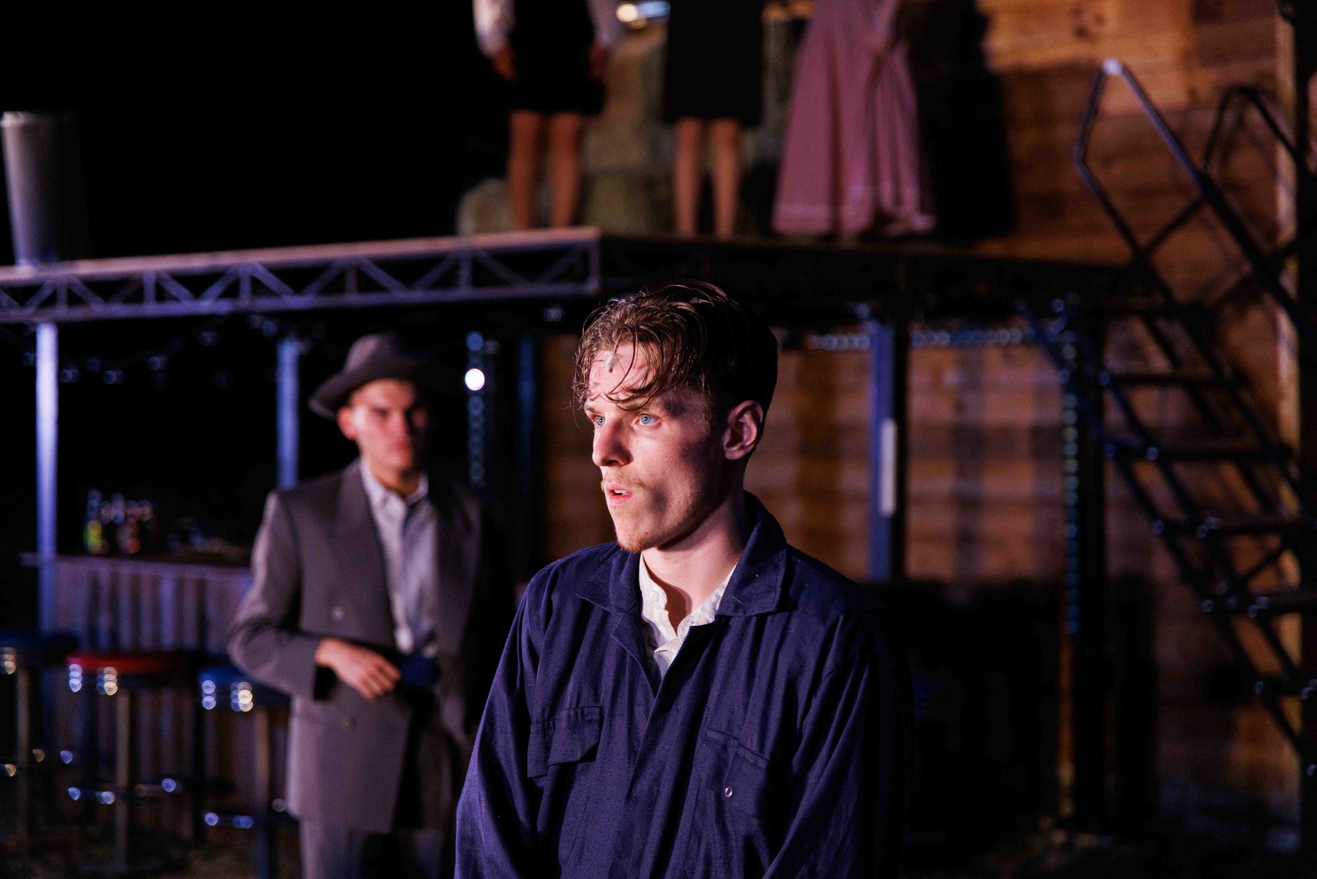 Two male performers in 1930s suits and workwear stand in front of a scaffold set, with ensemble members on the platform above.