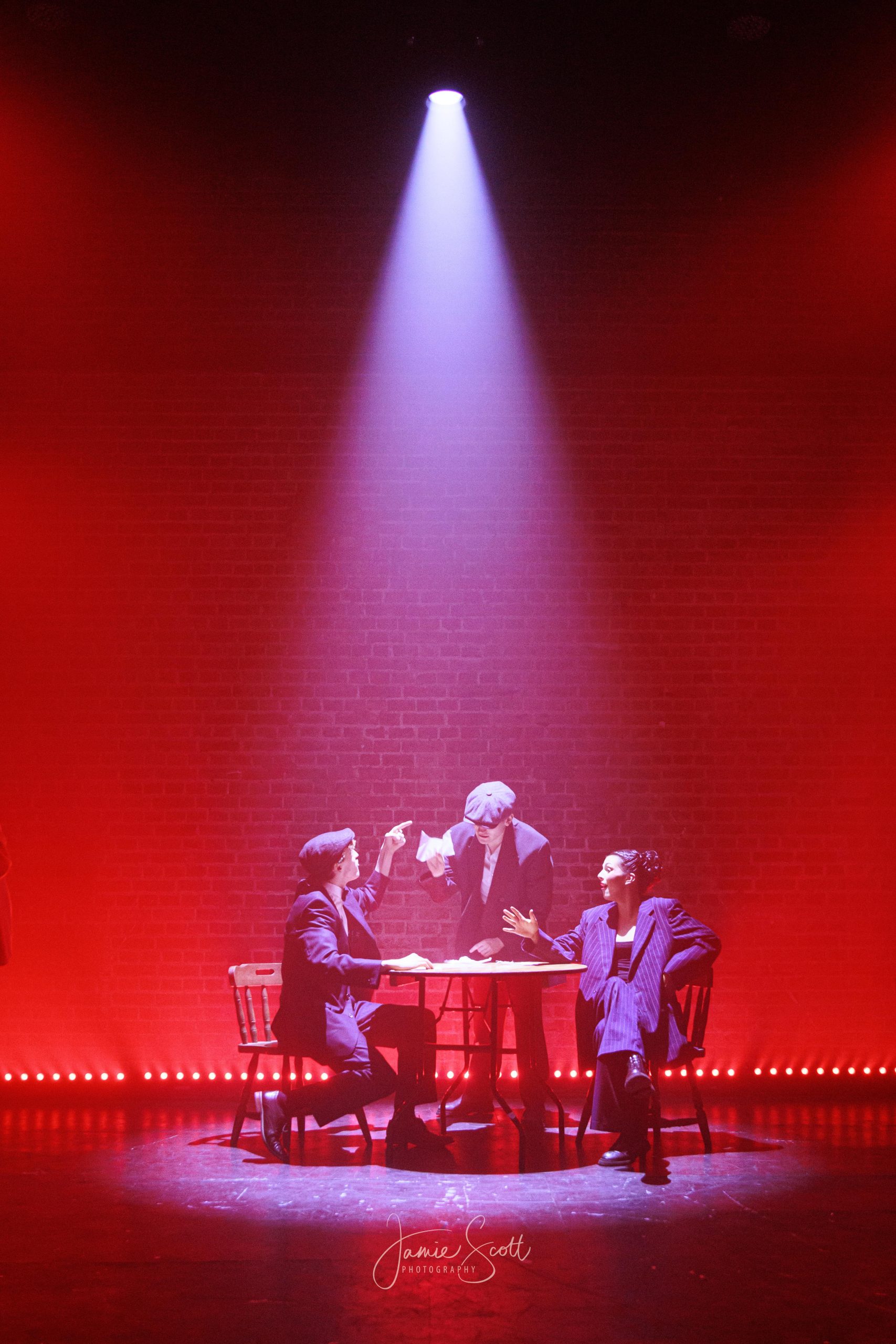 Performers sit around a round table under a single white spotlight on a stage washed in red.