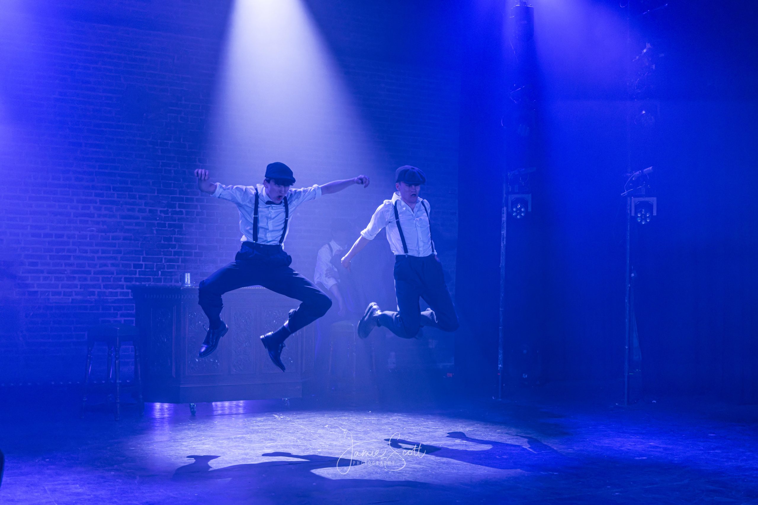 Two dancers in flat caps and braces leap in mid-air under blue spotlights against a brick wall.