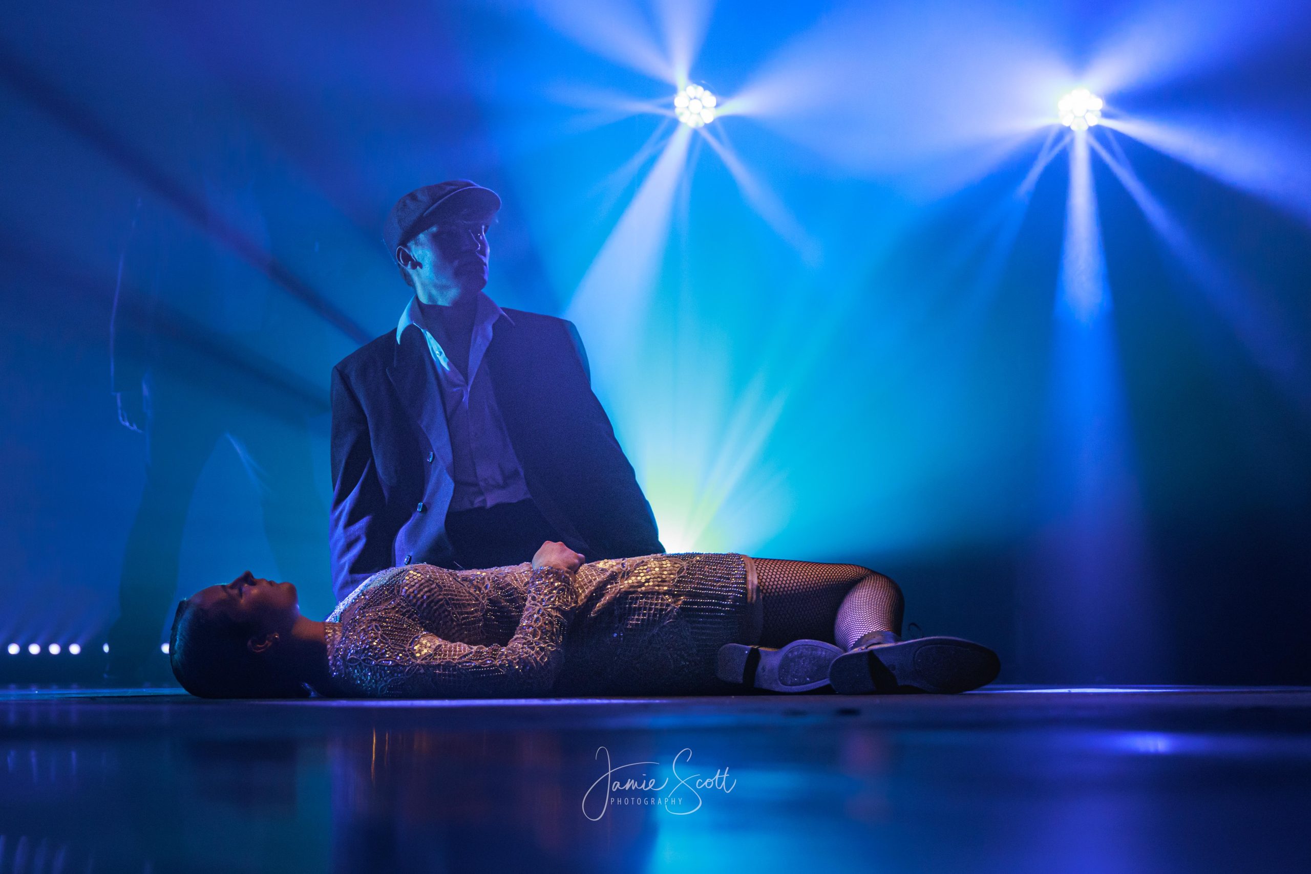A man in a flat cap and overcoat kneels over a woman lying on the stage in a sequinned dress, under blue beams.