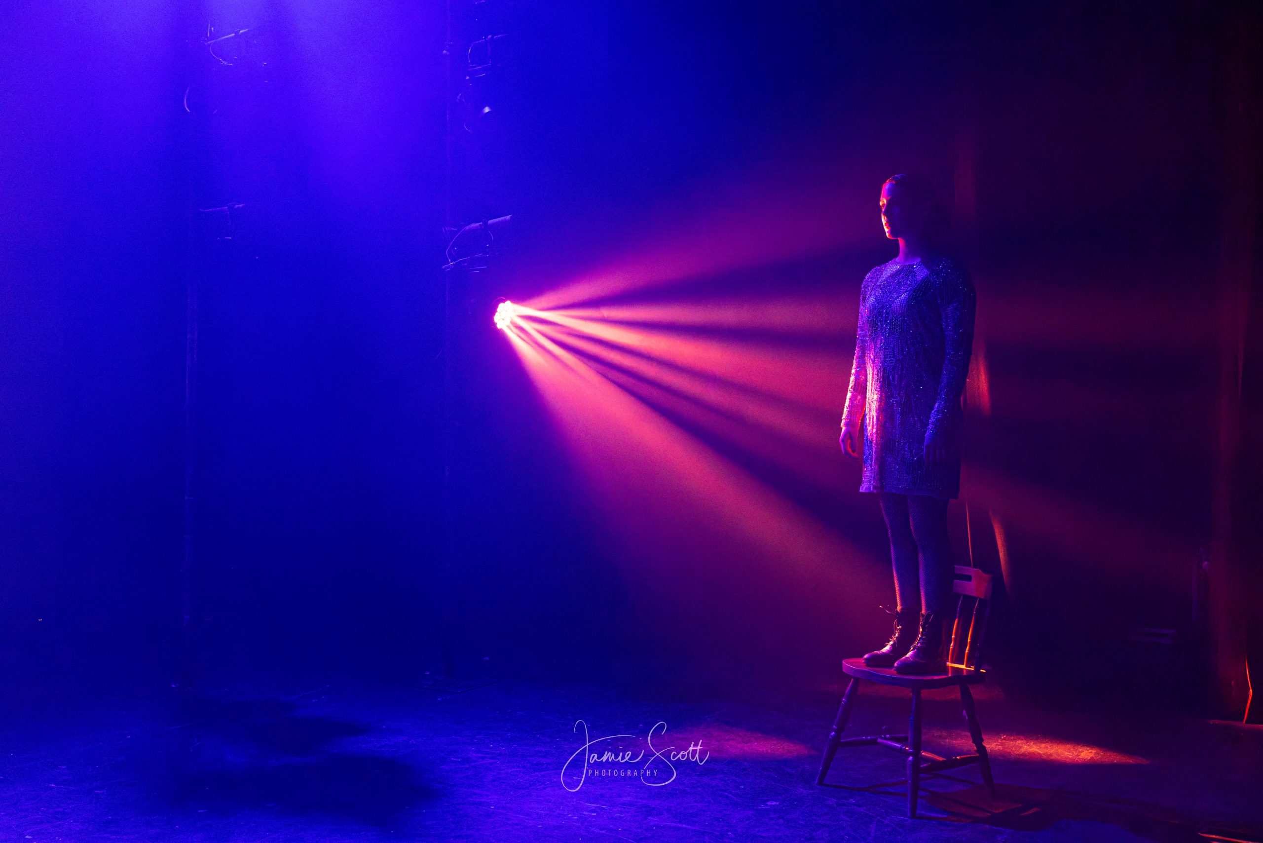 A performer stands on a chair on a dark blue stage, crossed by a single red beam of light.