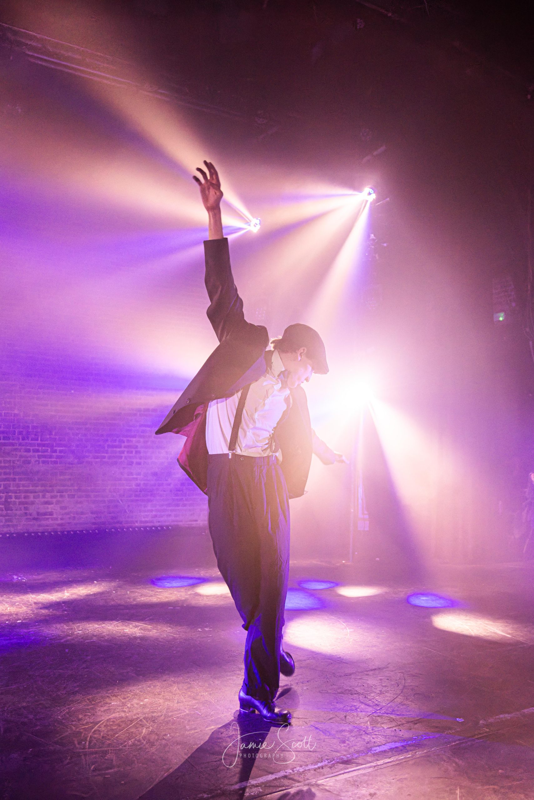 A dancer in a flat cap and suit stretches one arm upward in a haze of pink and white backlight.