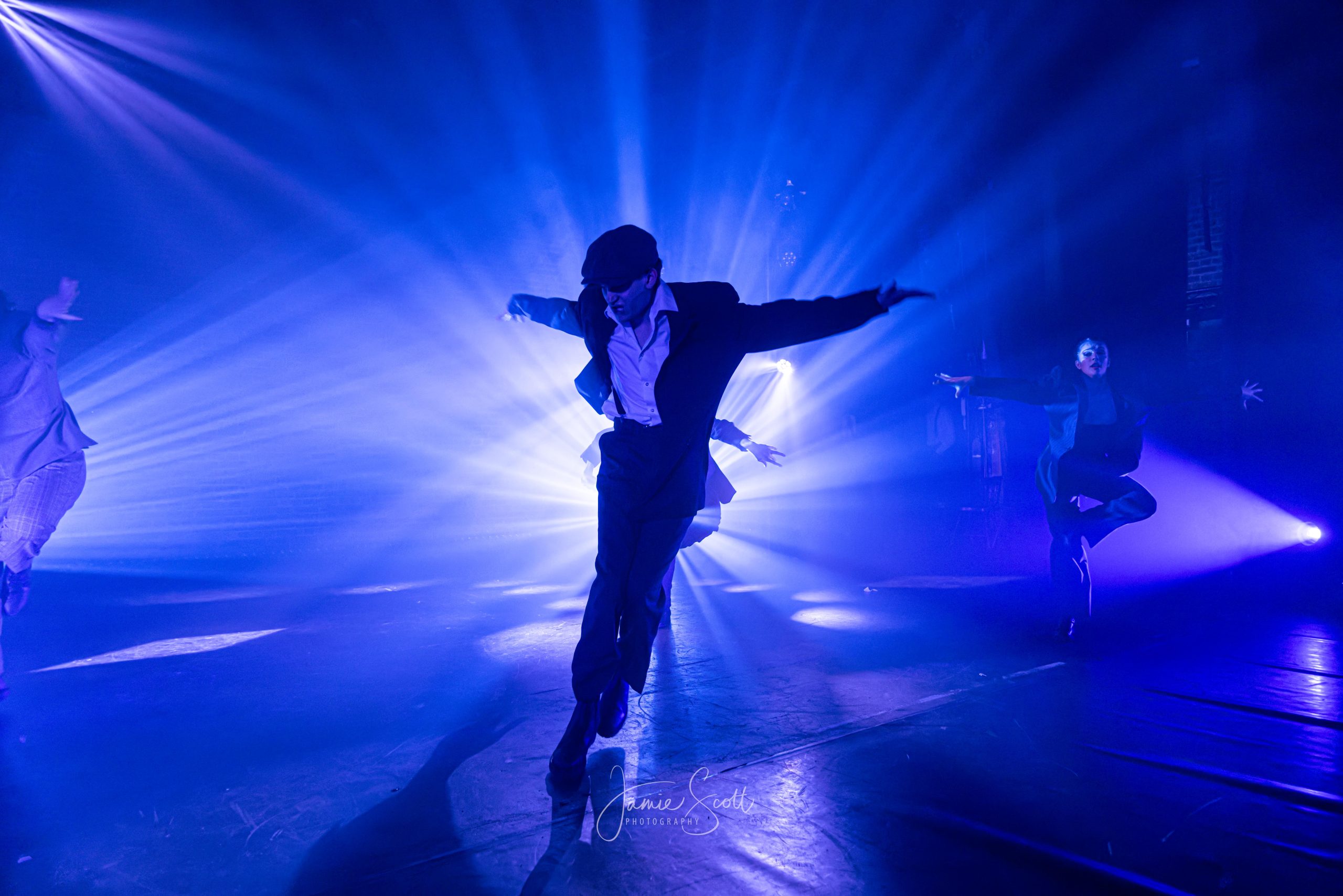 A dancer in a flat cap and suit performs centre stage, backlit by bright blue beams, with dancers either side.
