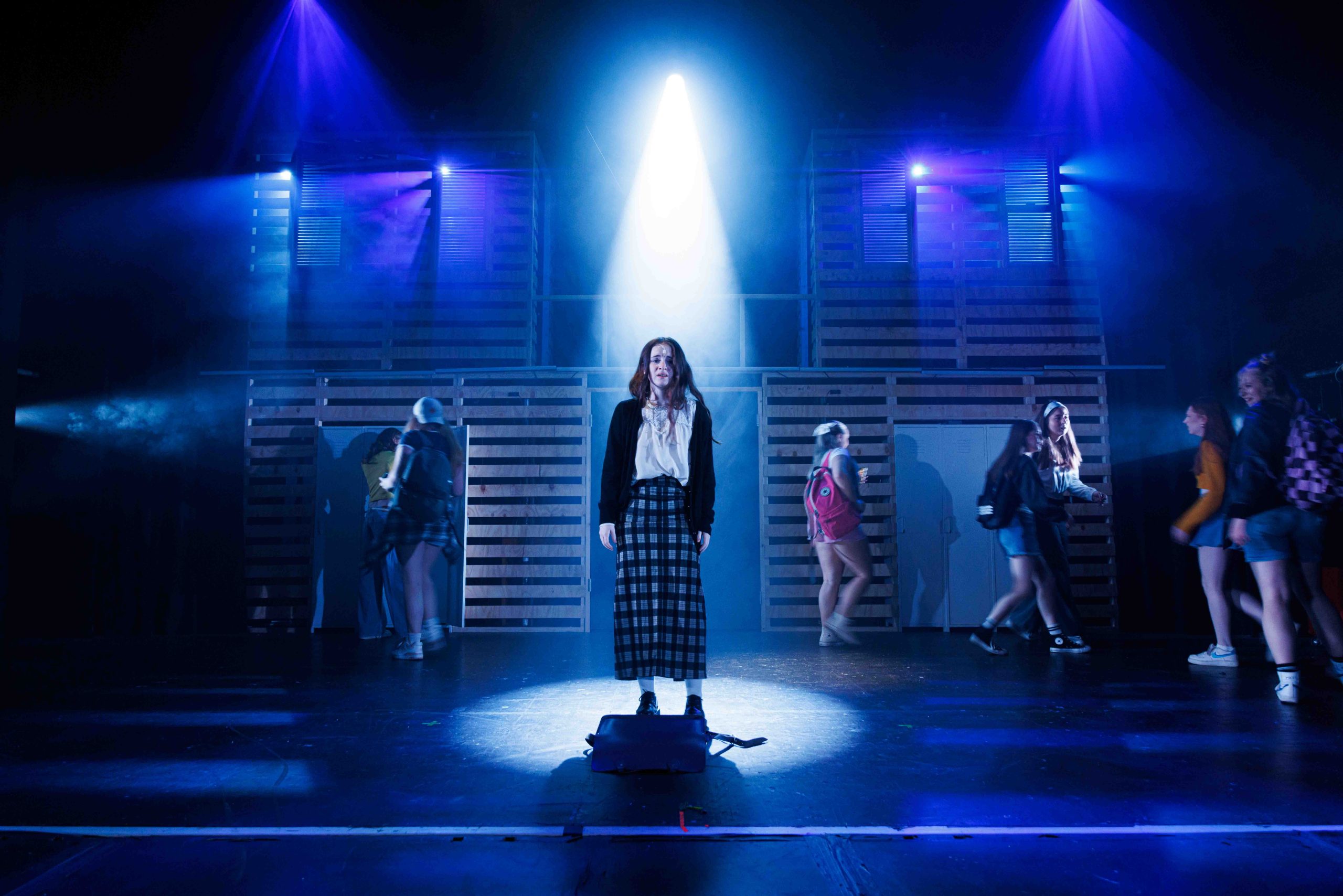 A girl in a long plaid skirt stands alone in a pool of light while classmates move past her.