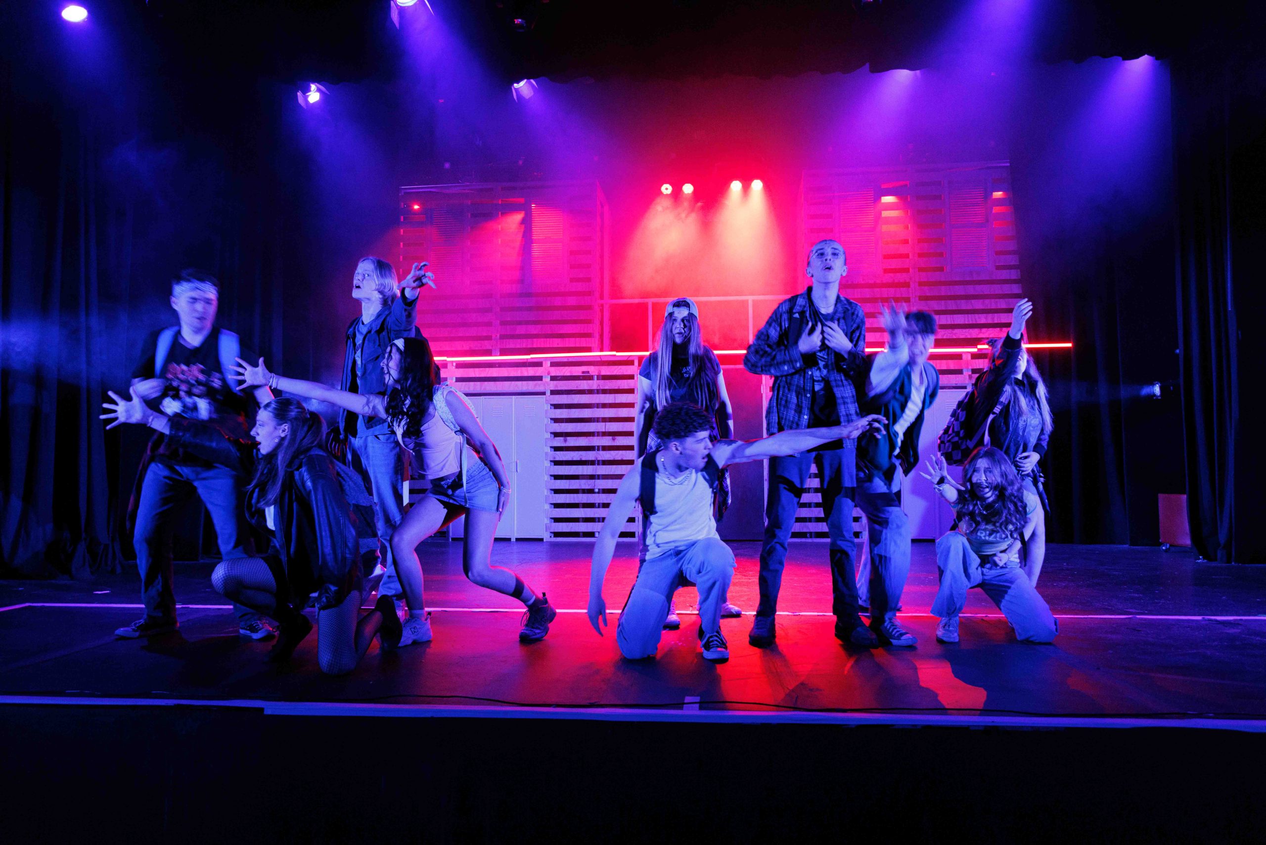 A large ensemble in casual high-school costume strike a dance pose under red and purple lights.