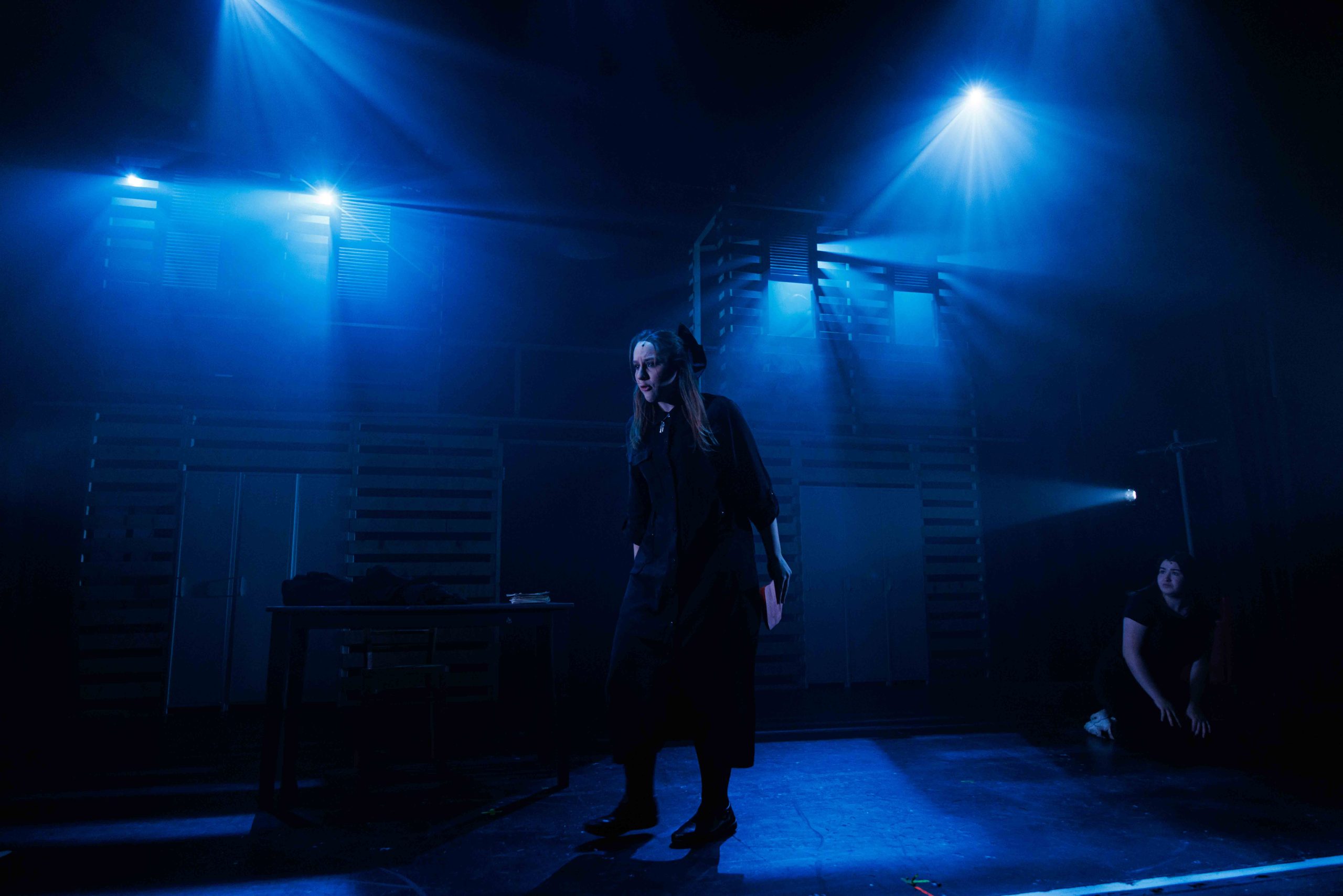 A young woman stands alone on a dim stage under blue lights, with a slatted wooden set behind.