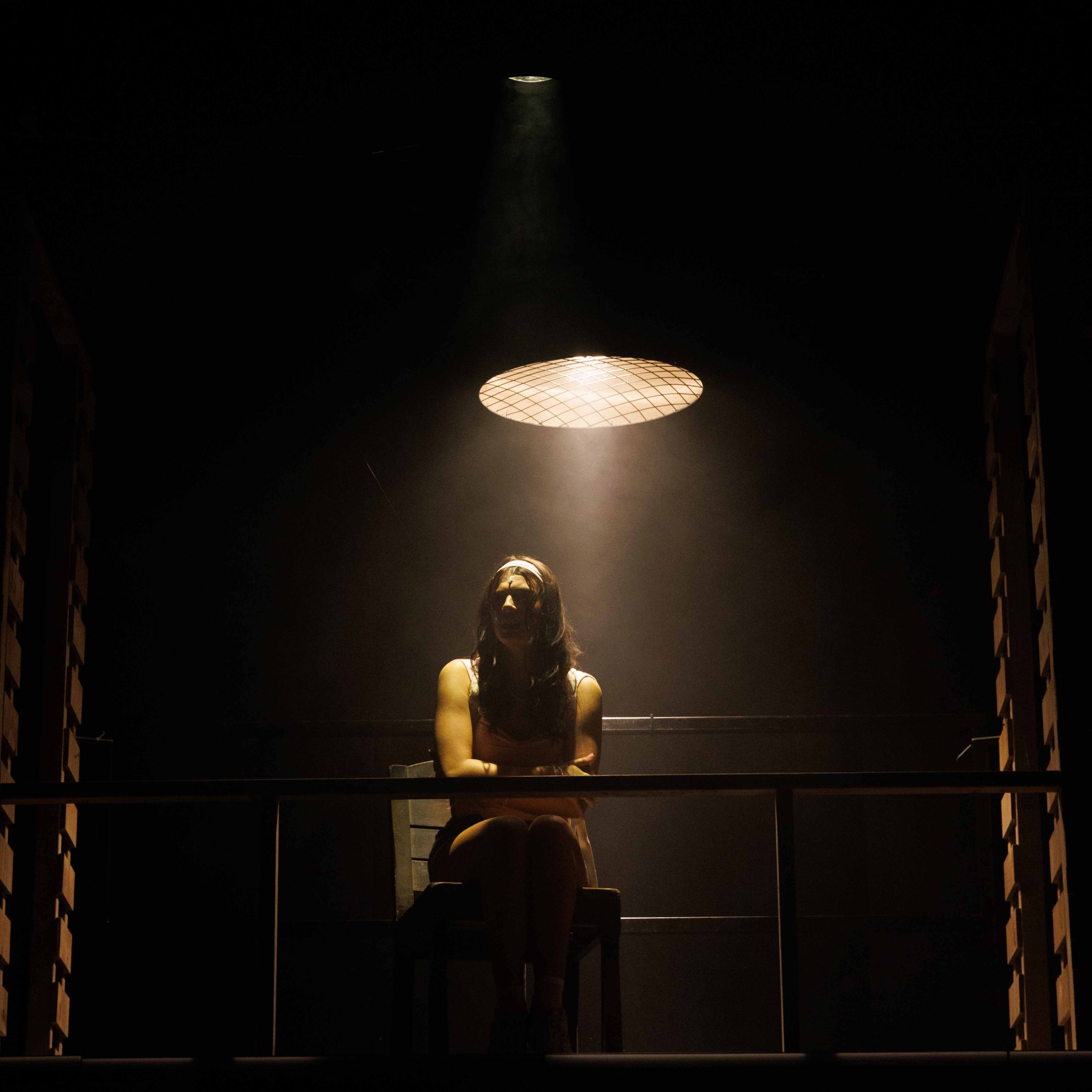A young woman sits alone beneath a single hanging lamp in a dark room.