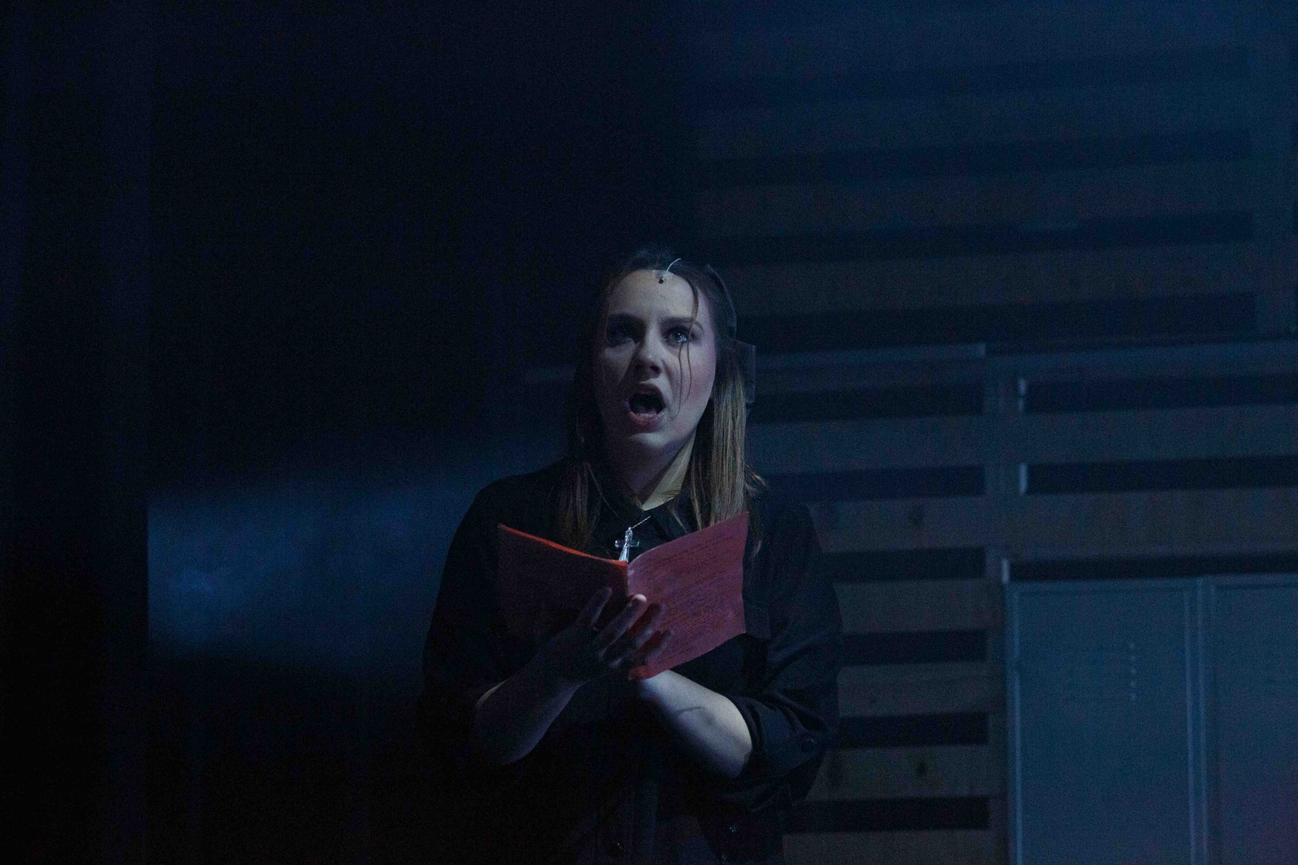 A performer holding an open red book sings in dim, cold blue light.
