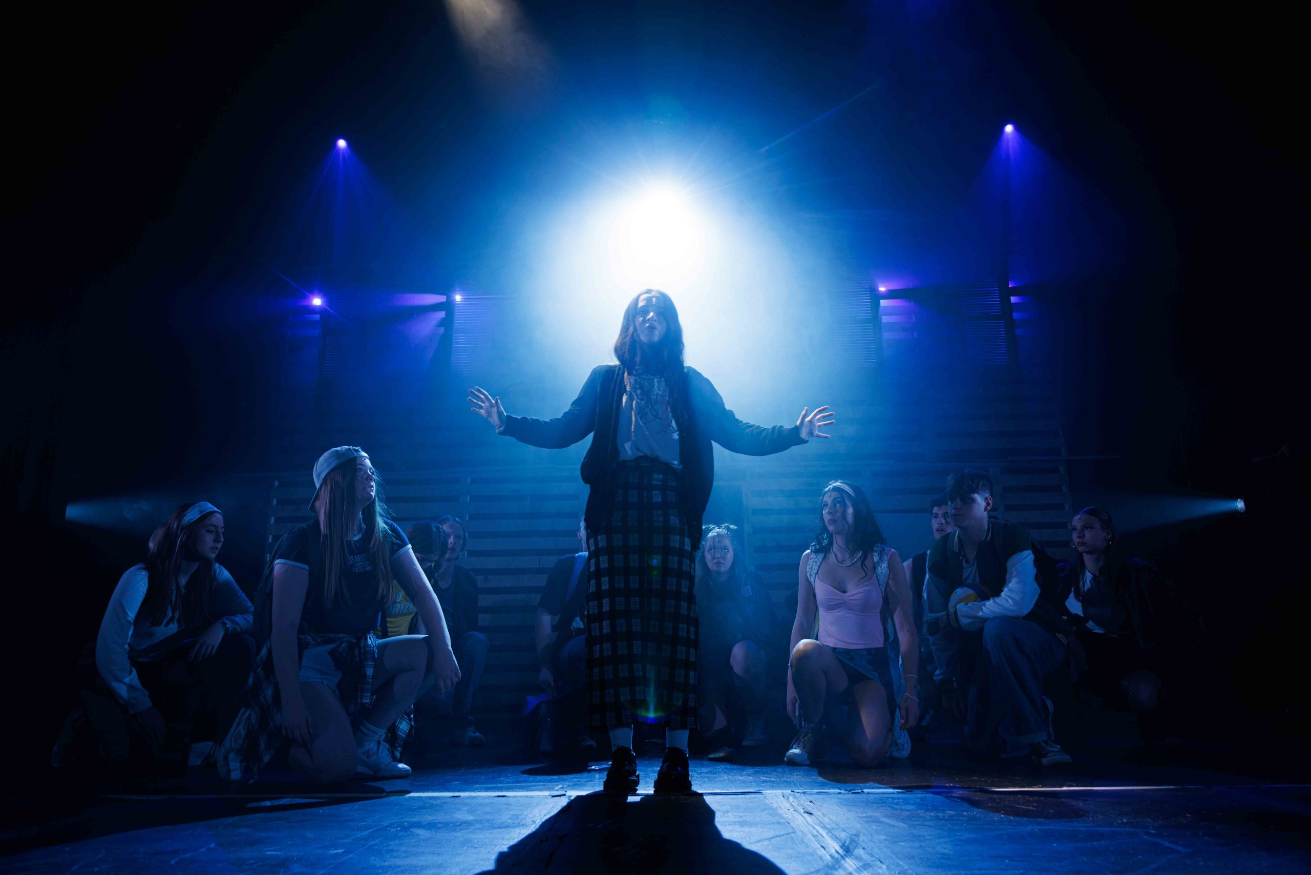 A girl in a long plaid skirt stands with arms open in a beam of blue light, with the ensemble crouched around her.