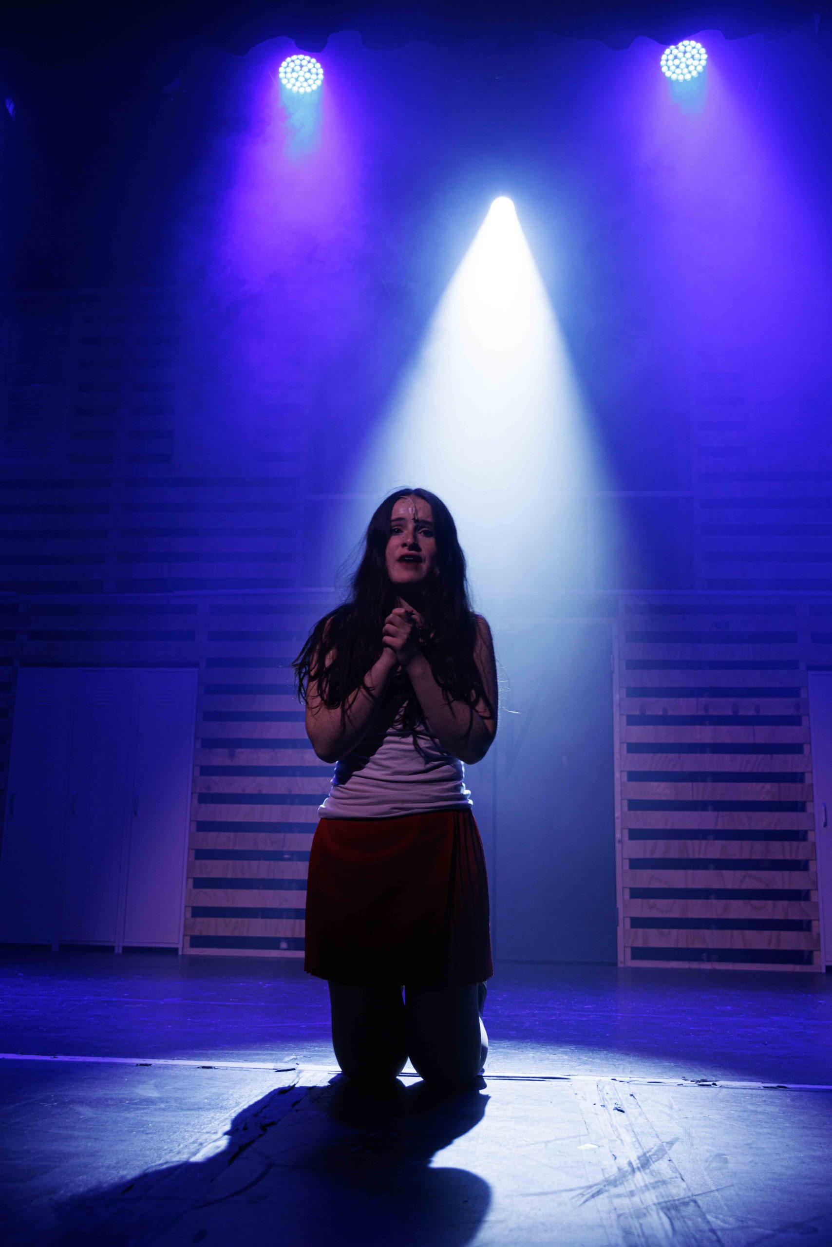 A young woman in a white top clutches her hands to her chest under a single white spotlight.