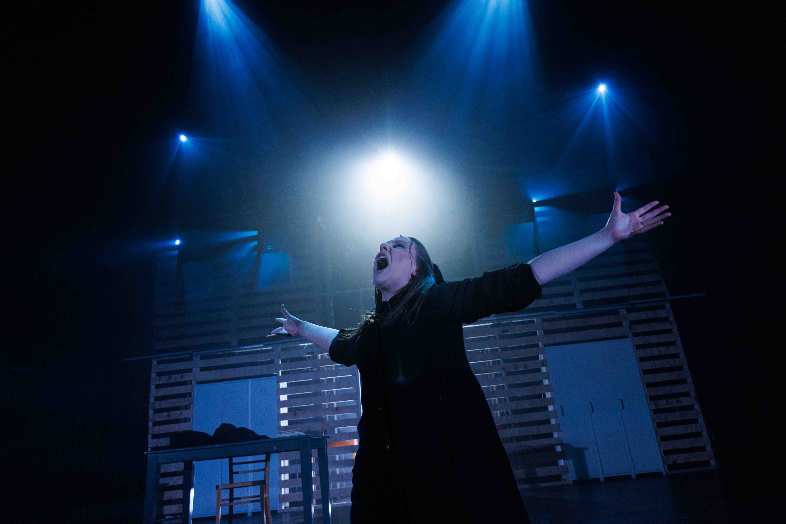 A performer in black sings with arms flung wide under blue spotlights.