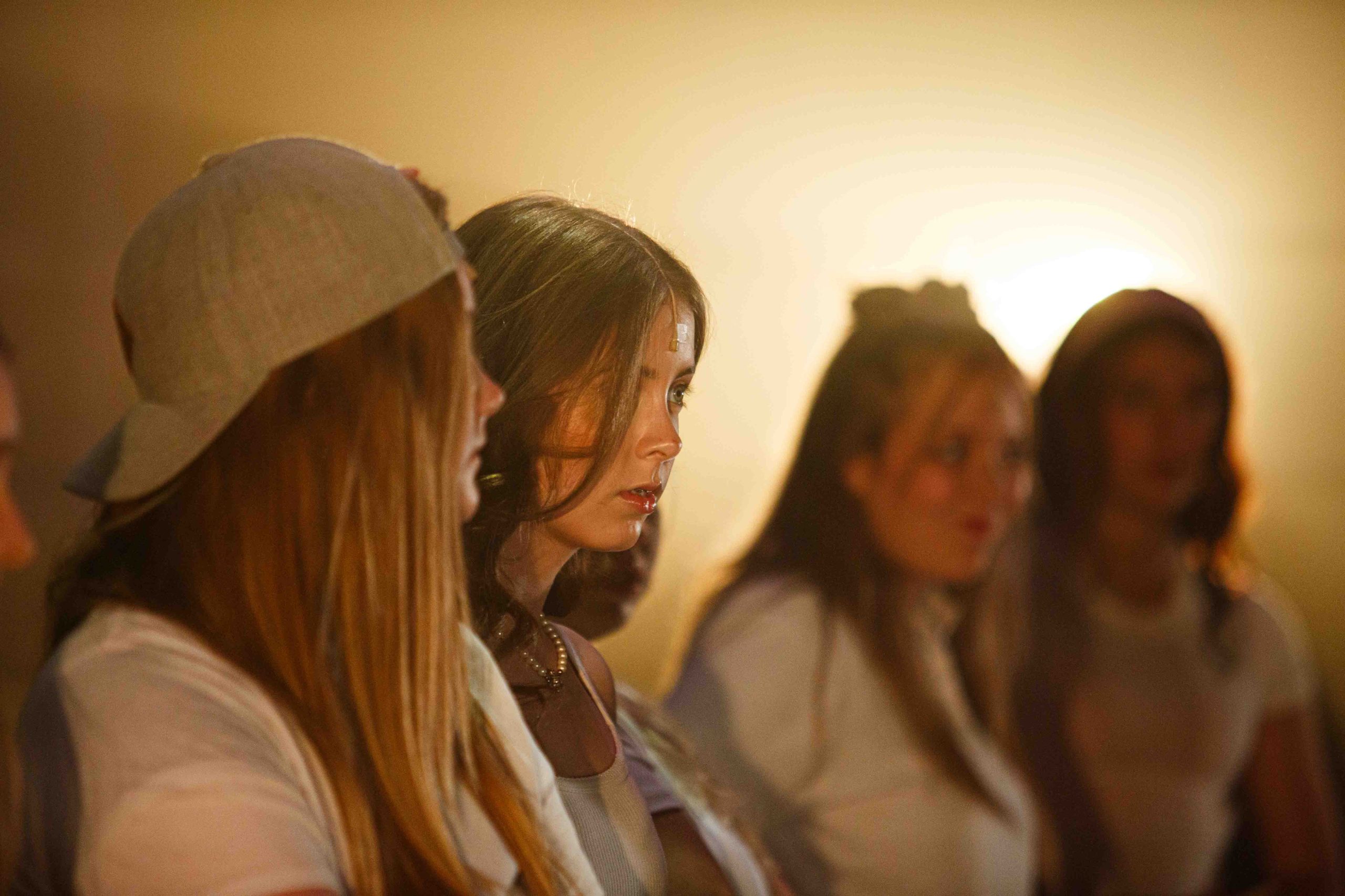 Close-up of four young women in white tops, one in a baseball cap, looking to the side in warm golden light.