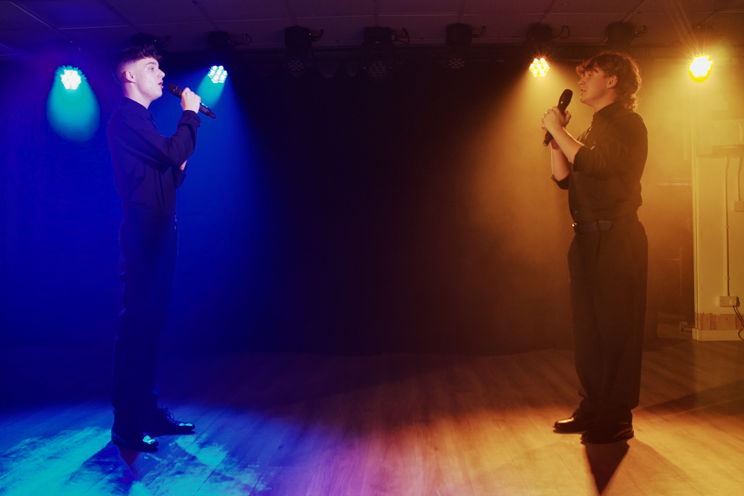 Two singers with microphones face each other across a stage split by blue and amber light.