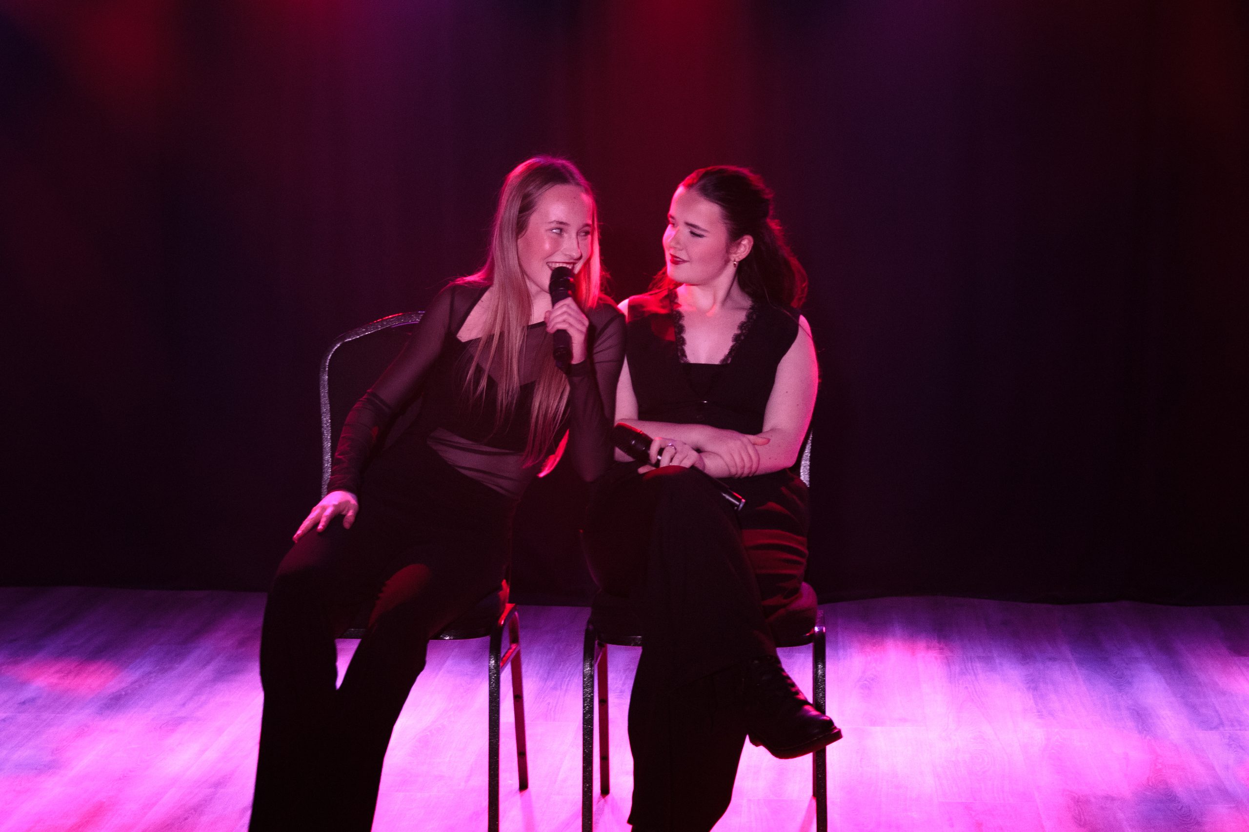 Two young women sit side by side on chairs, smiling at each other, on a pink-lit stage.