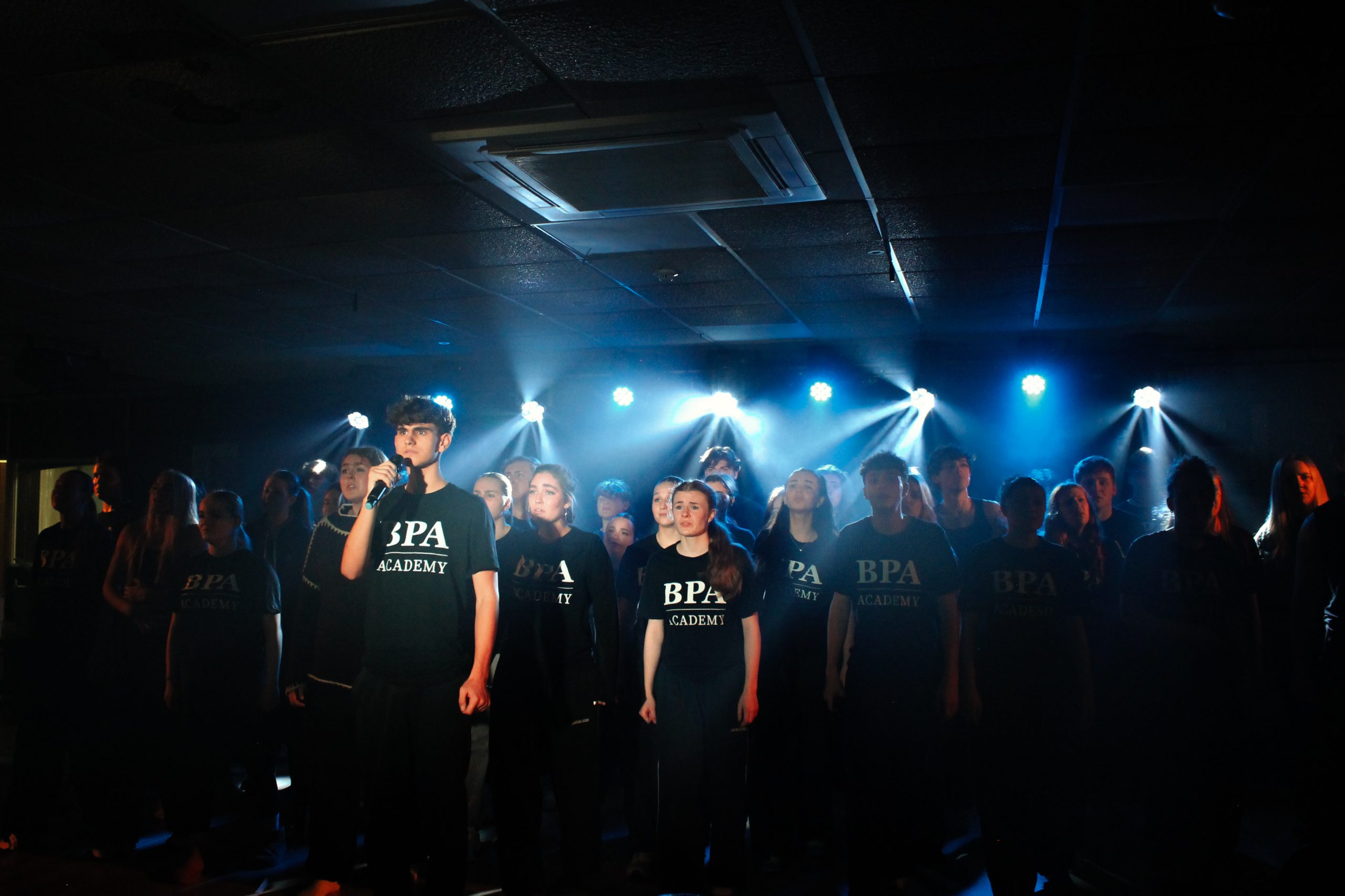 The company in black BPA Academy T-shirts stand together on stage under blue lights.