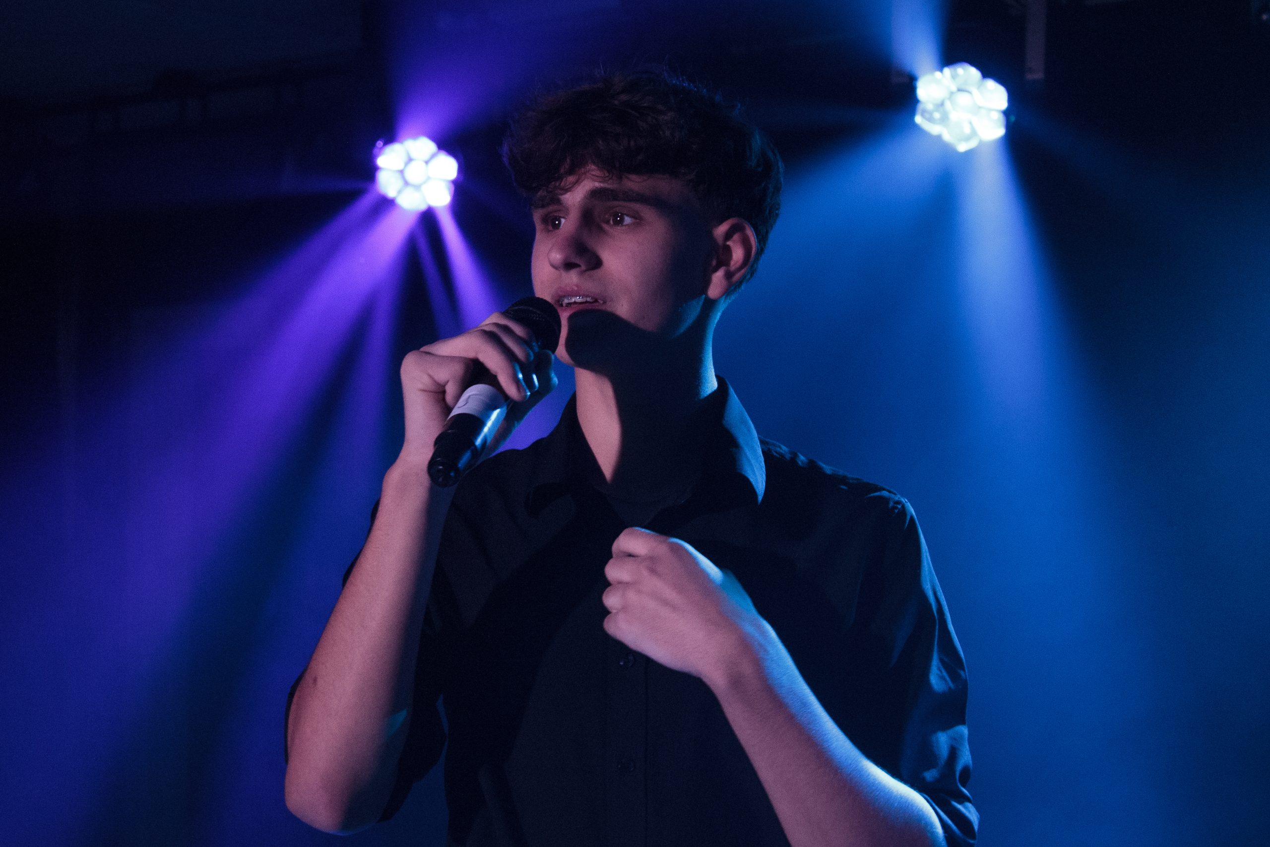A young man sings into a microphone with his hand on his chest under blue spotlights.