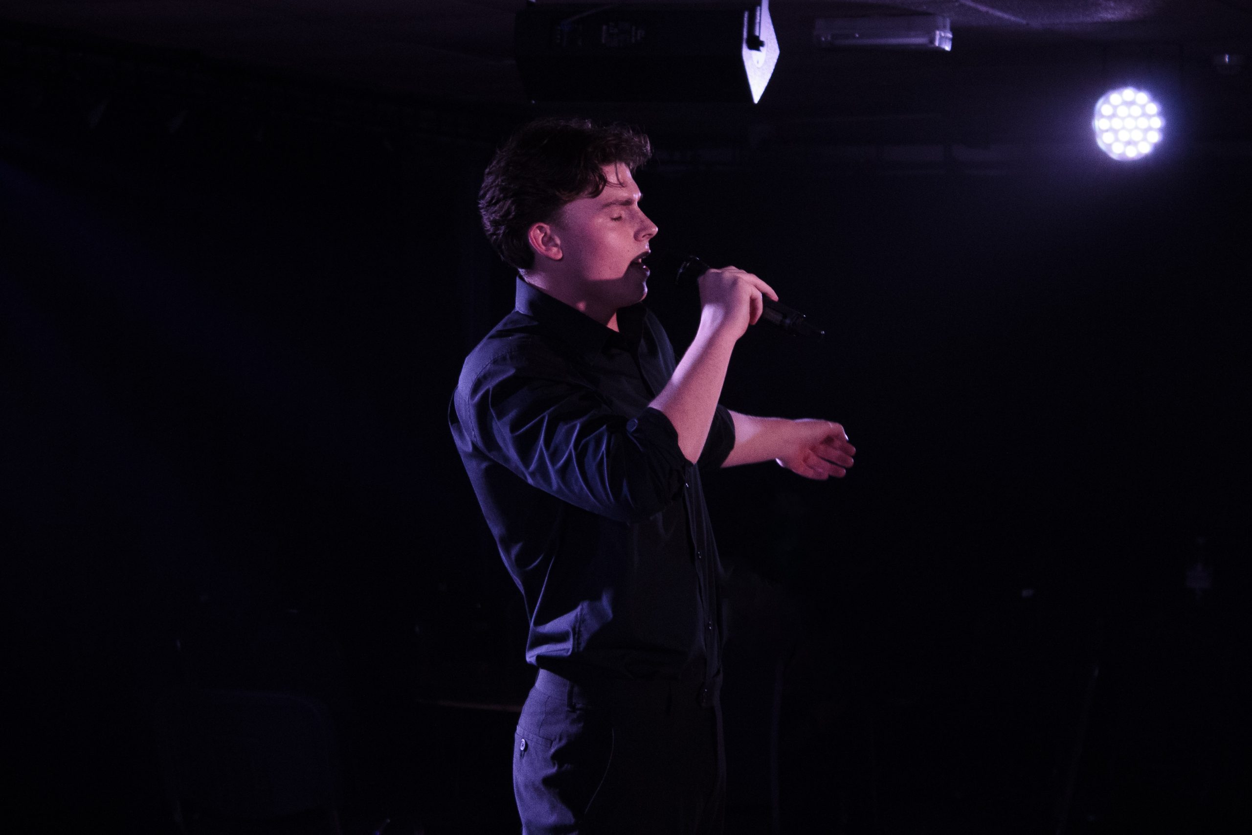 A young man in black sings into a microphone, gesturing with one hand, on a dark stage.