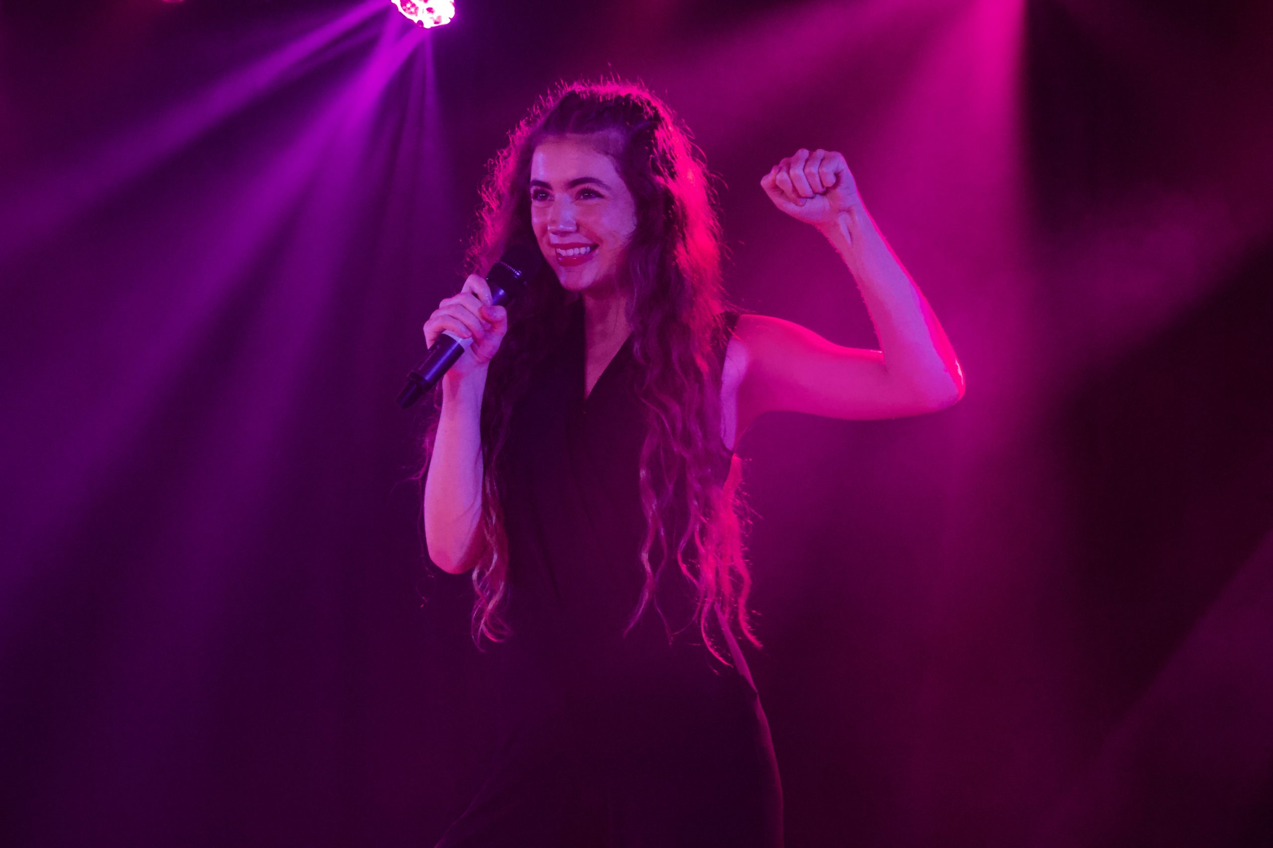 A singer with long curly hair raises a fist triumphantly, lit in magenta.