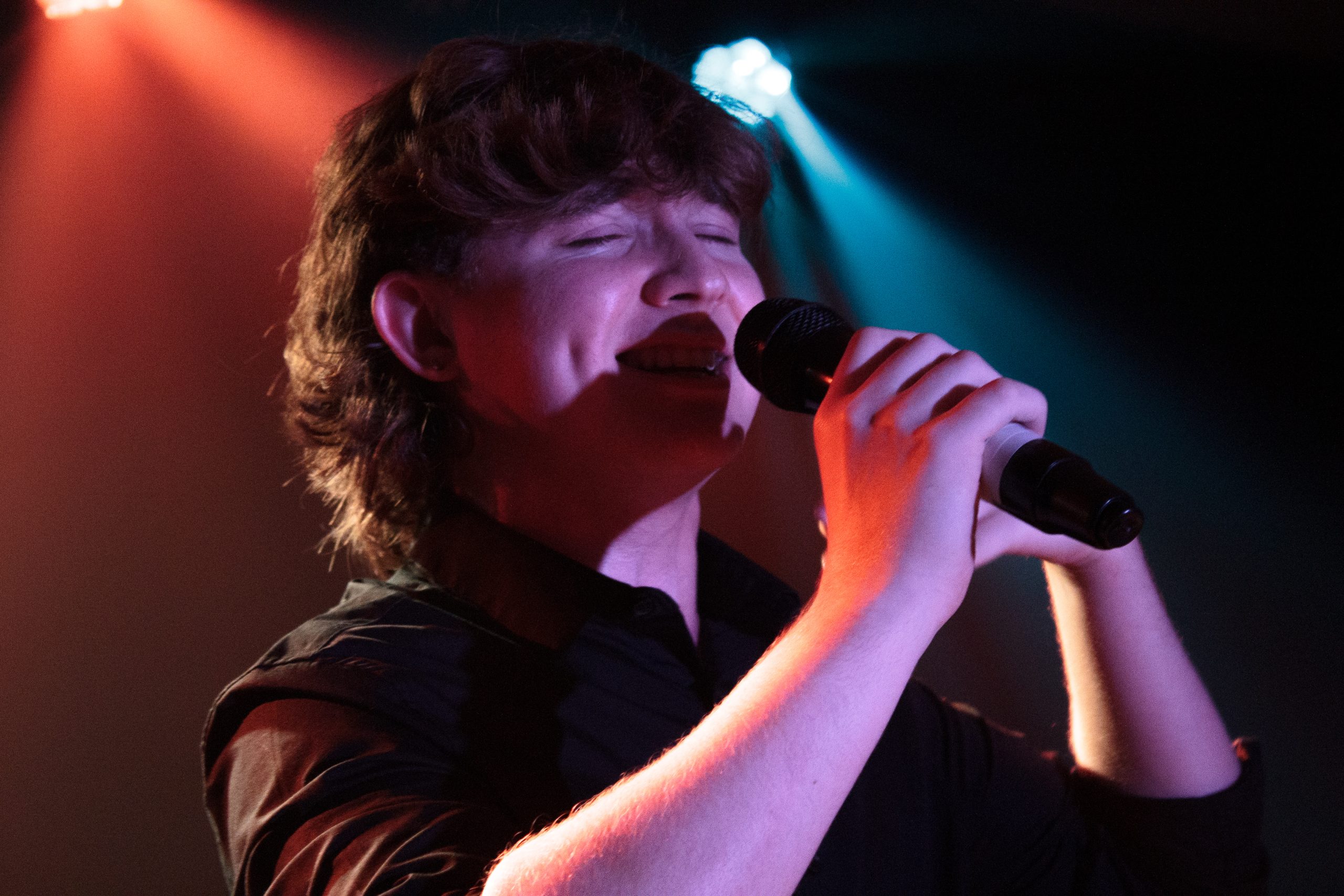 Close-up of a young singer with a microphone, eyes closed, lit in red and blue.
