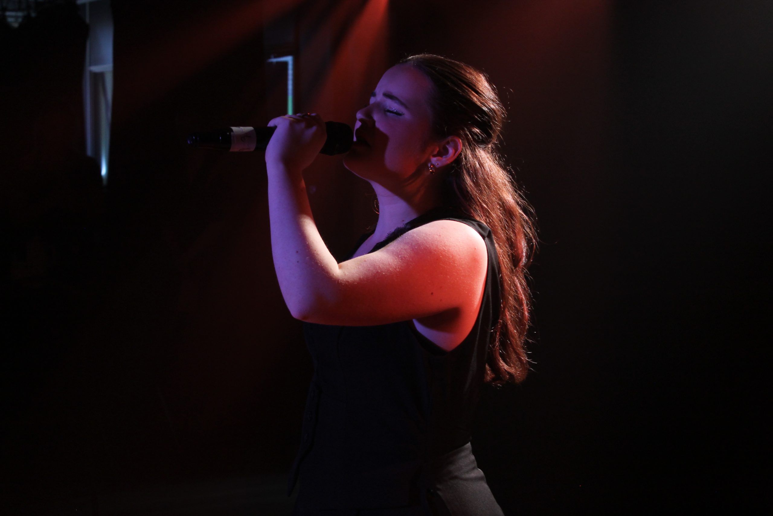 A young woman sings into a microphone with eyes closed, lit by warm red light.