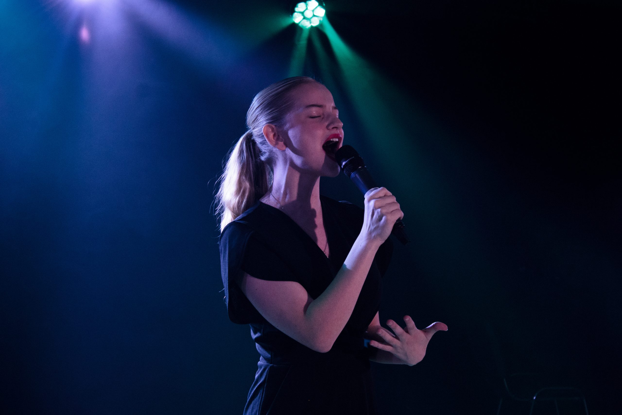 A young woman with a ponytail sings passionately into a microphone under teal light.