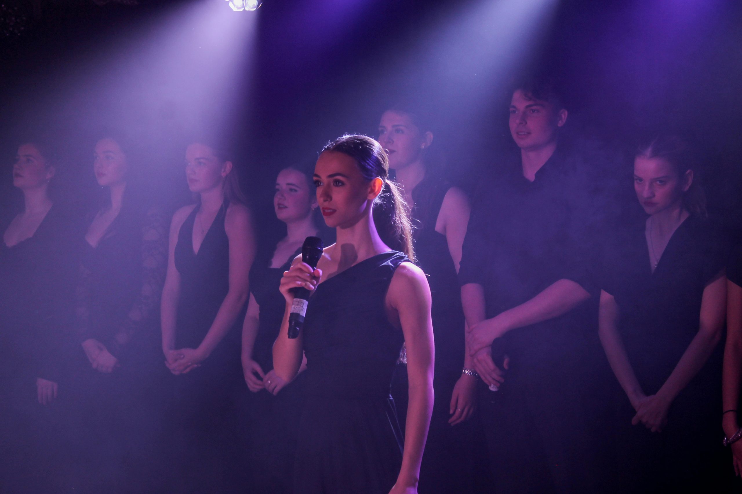 A young woman in a black dress sings into a microphone in front of the company in purple haze.