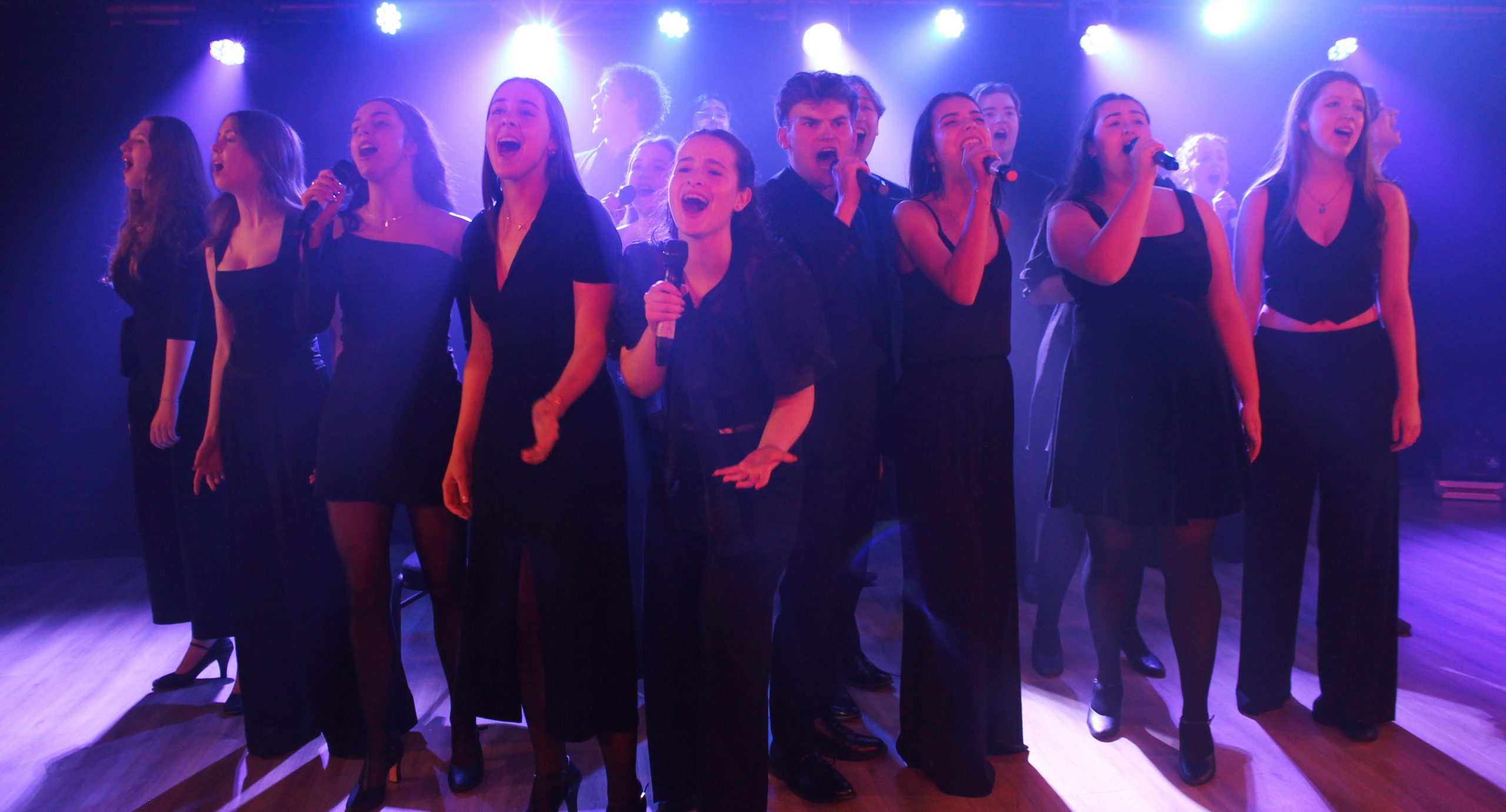 The company in black outfits sing together under purple lights.