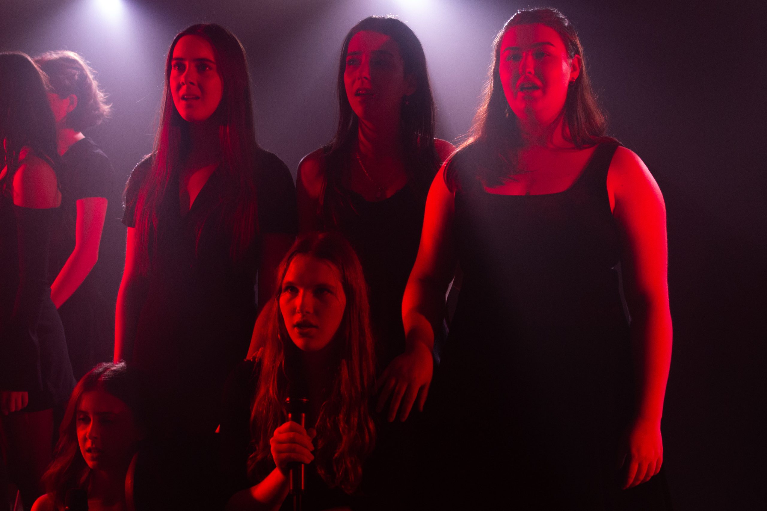 Three young women sing together in red light, with another performer kneeling in front.
