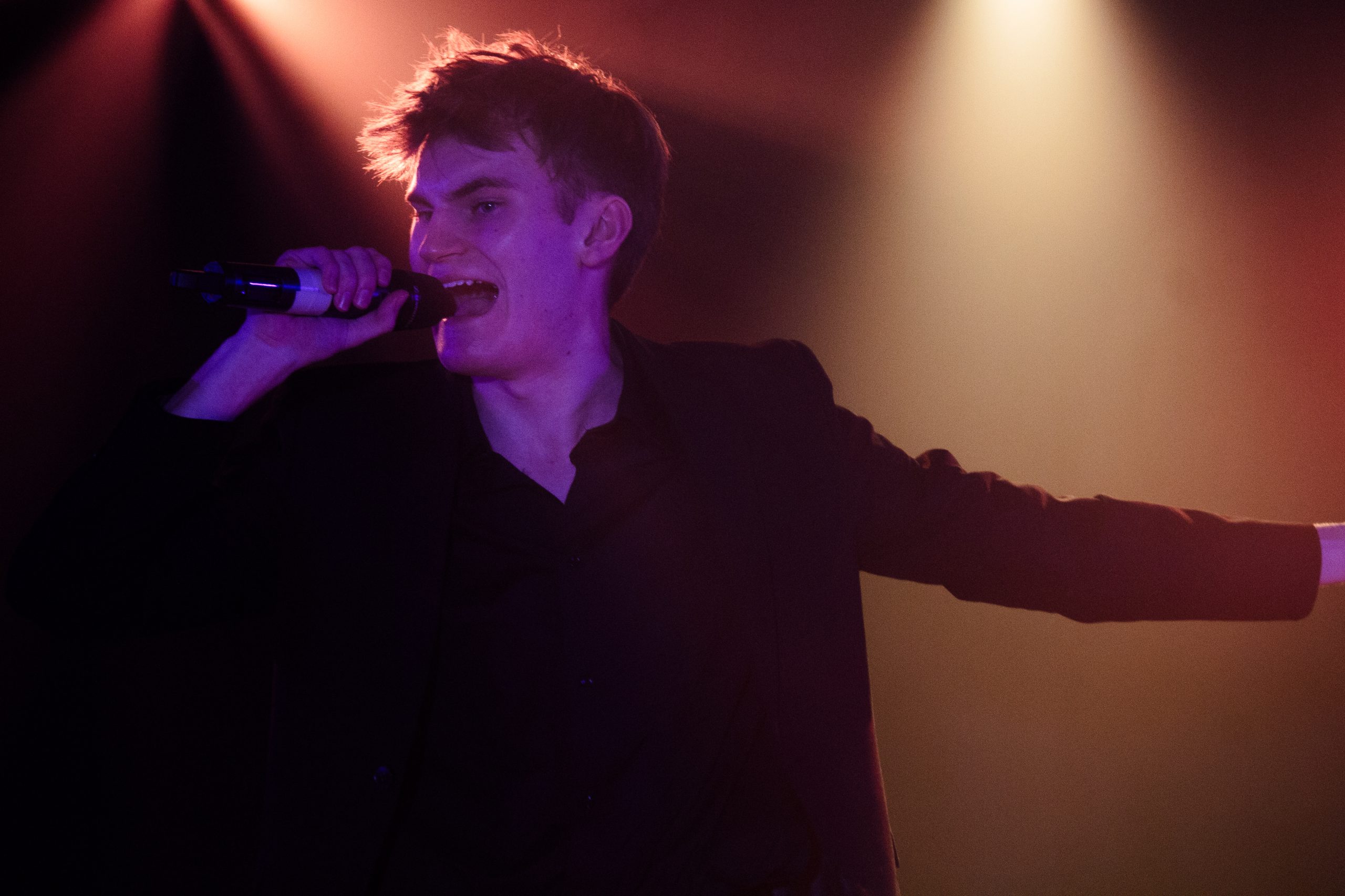 A young man sings passionately into a microphone with one arm outstretched.