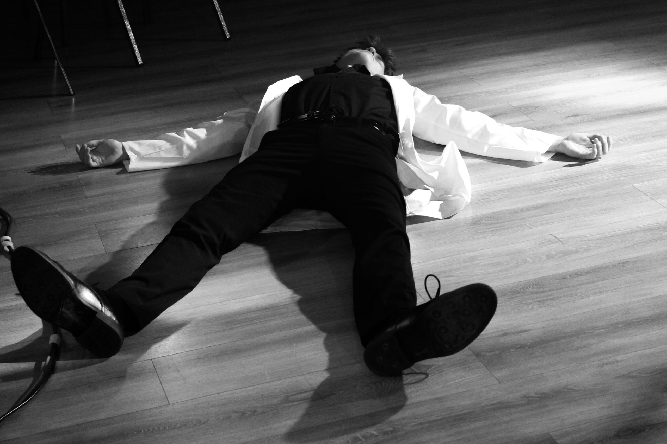 Black-and-white photo of a performer in a white jacket lying spread-eagled on a wooden floor.