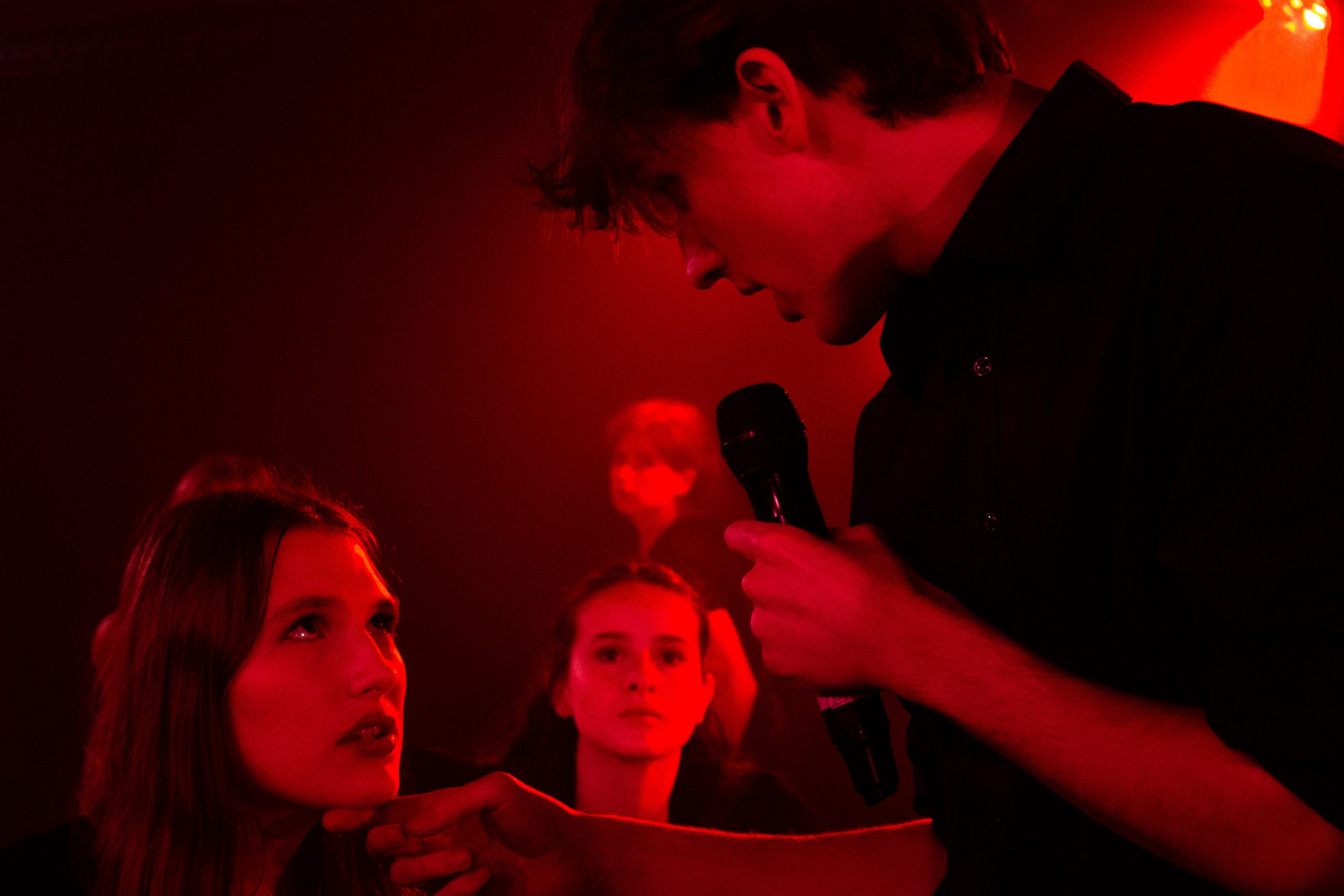 A young man leans in to sing to a young woman in a red-lit scene.