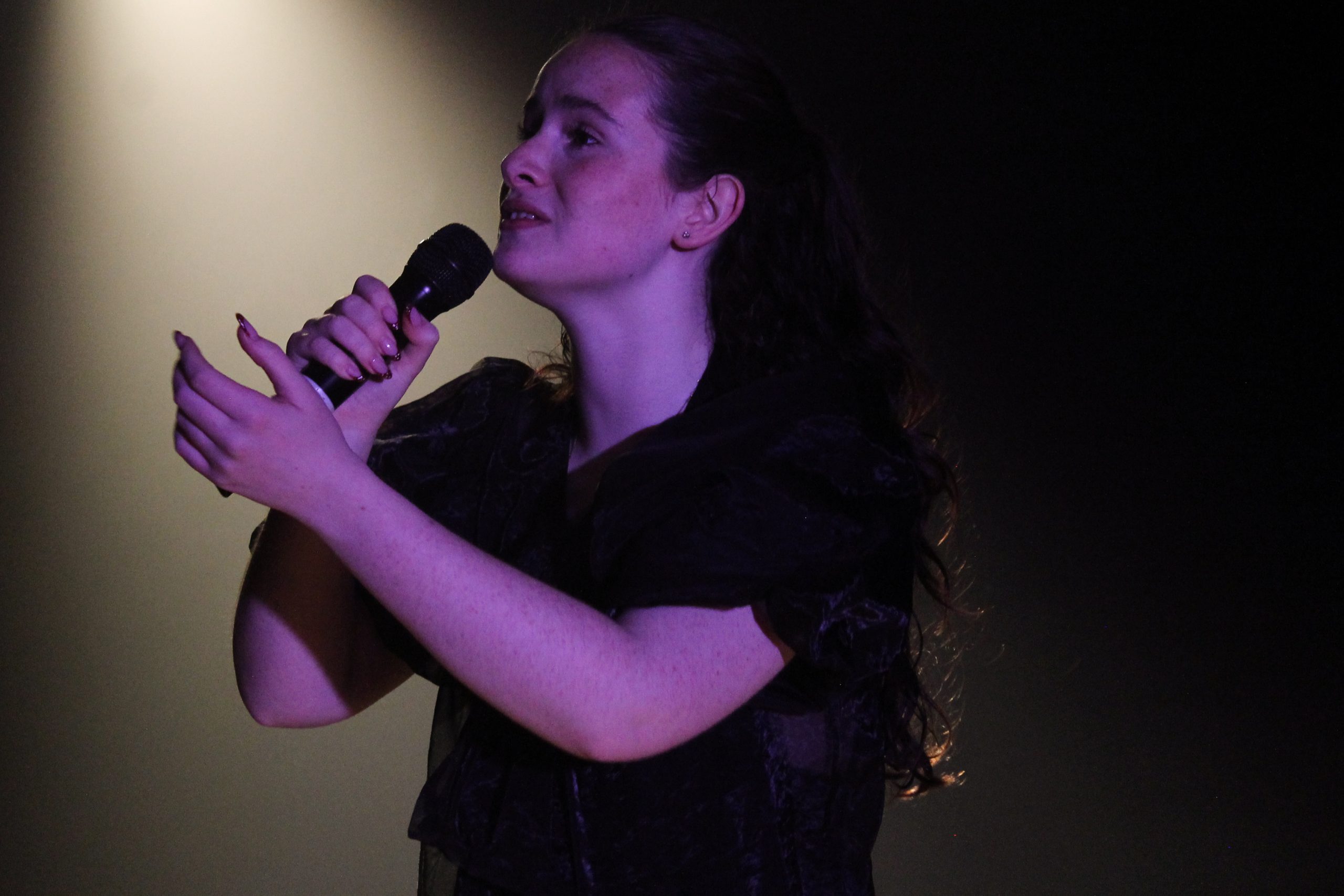 A young woman holds a microphone with both hands, lit by a single warm spotlight.