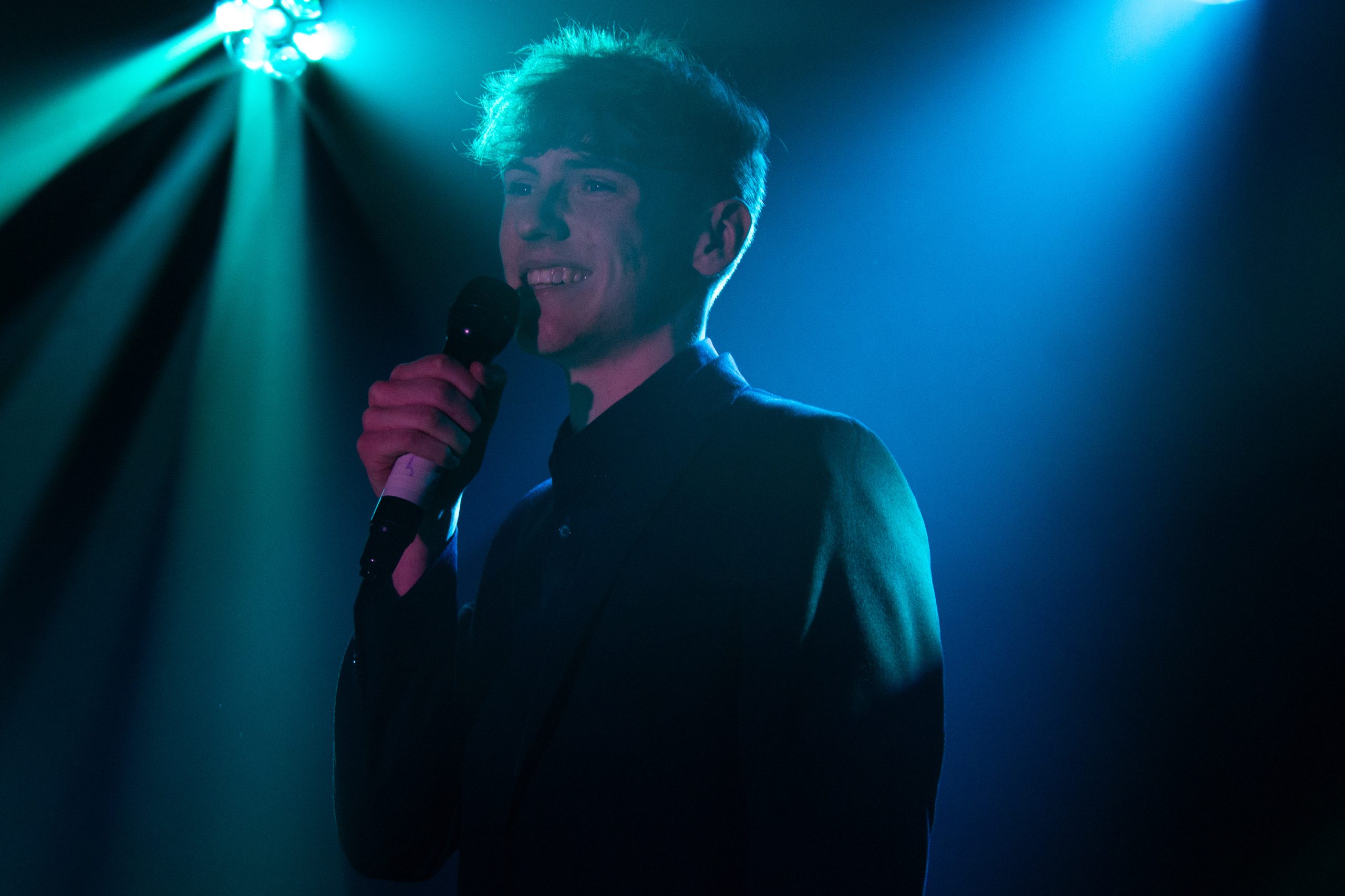 A smiling young man sings into a microphone under teal stage lights.