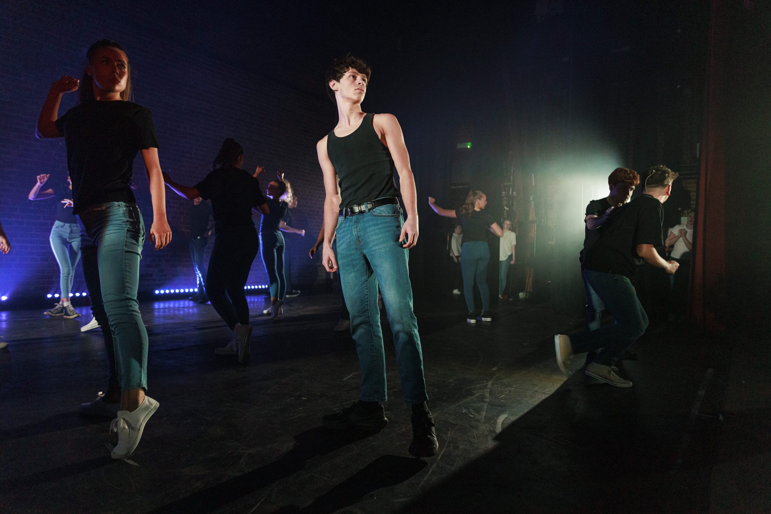 Dancers in jeans and dark tops perform in haze lit from the side.