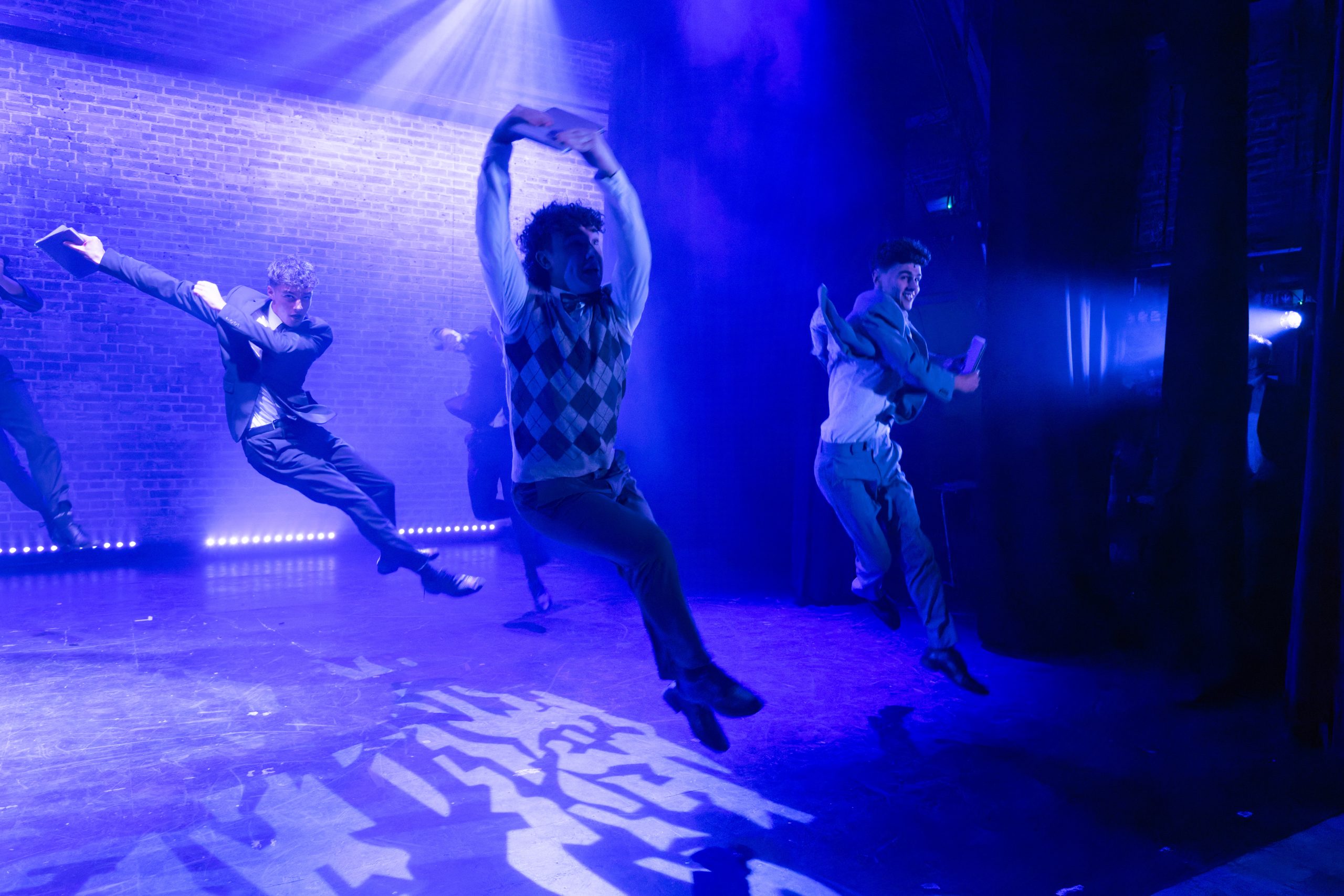 Three dancers leap and twist across a blue-lit stage with patterned light on the floor.