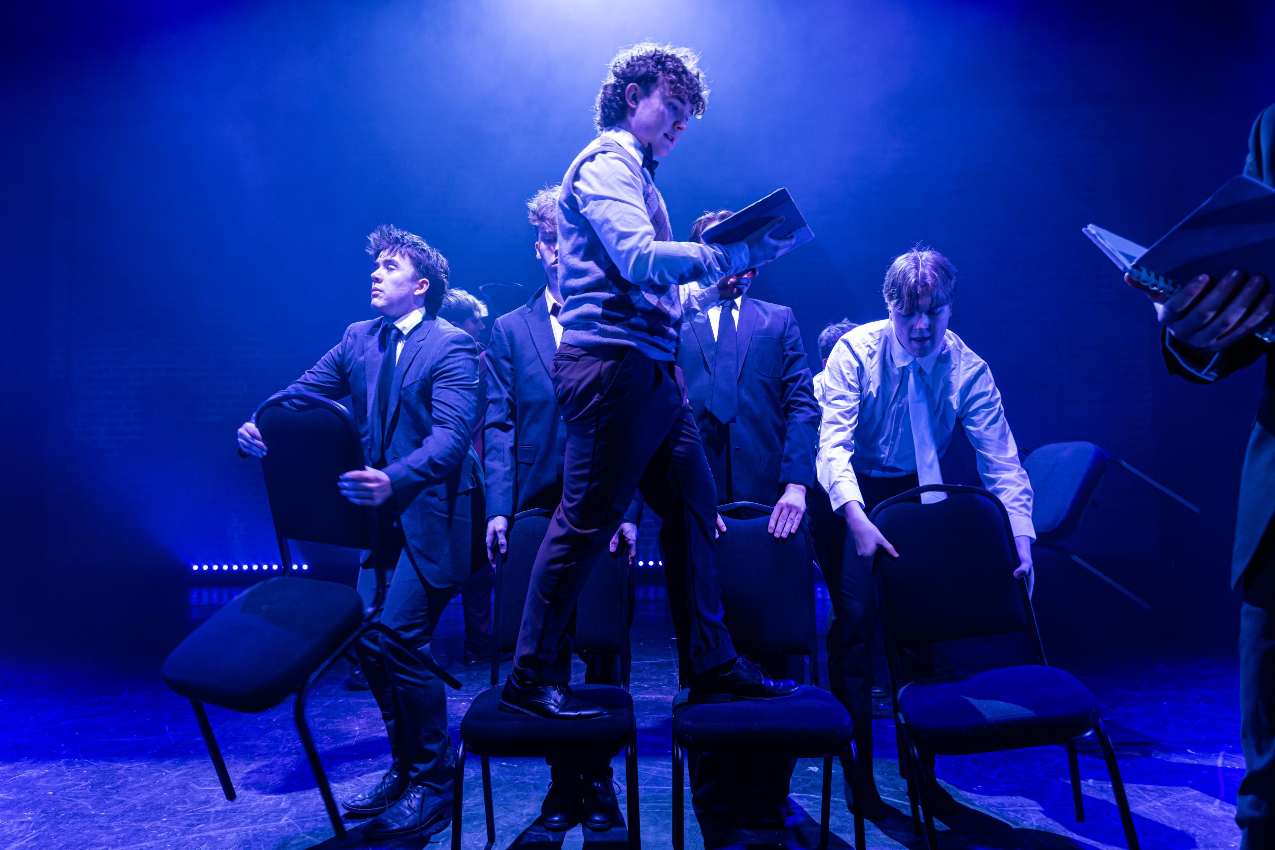 Performers in suits move between rows of chairs under blue light, one standing on a chair holding a book.