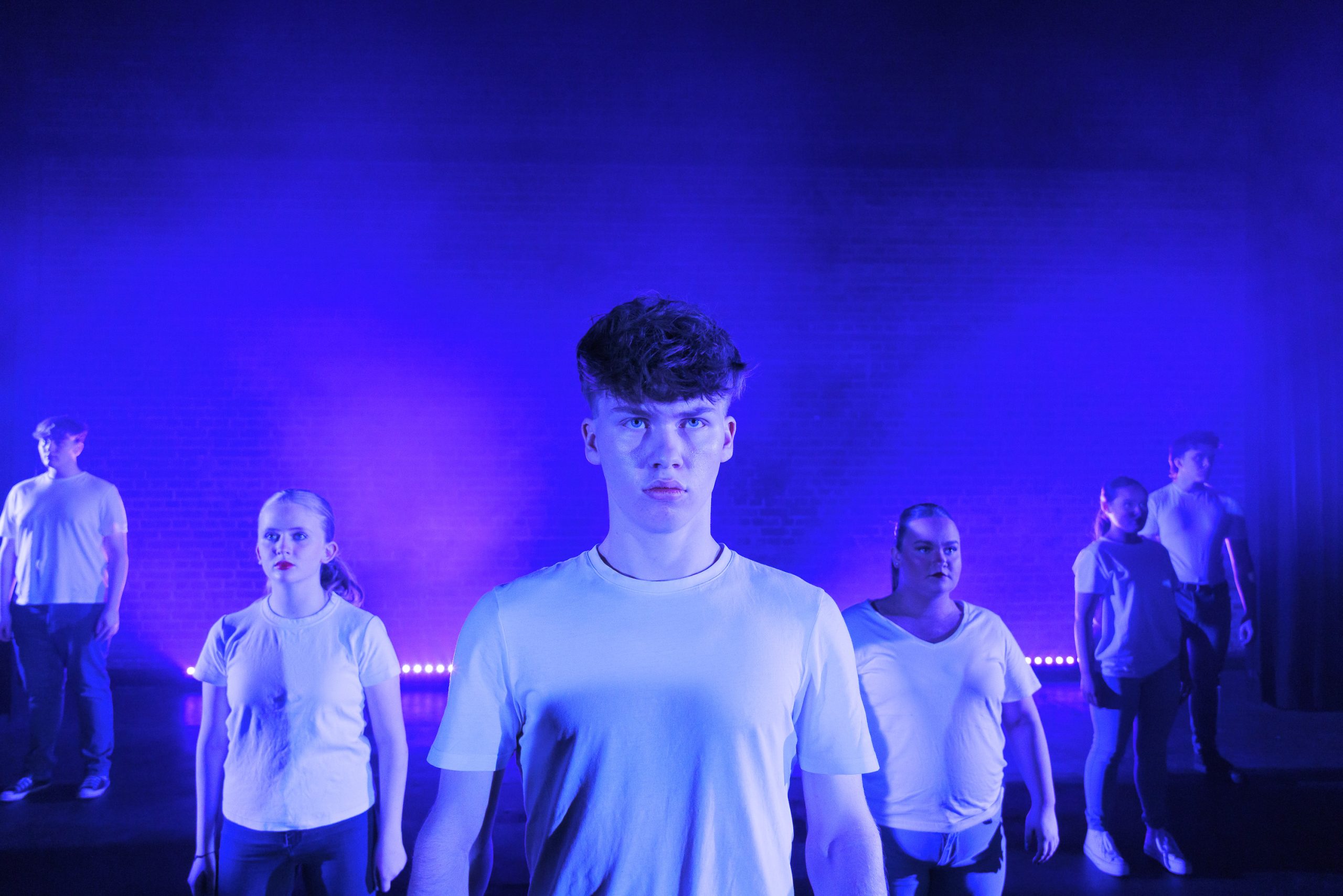 A young man in a white T-shirt looks straight ahead, with other performers in white behind him under blue light.