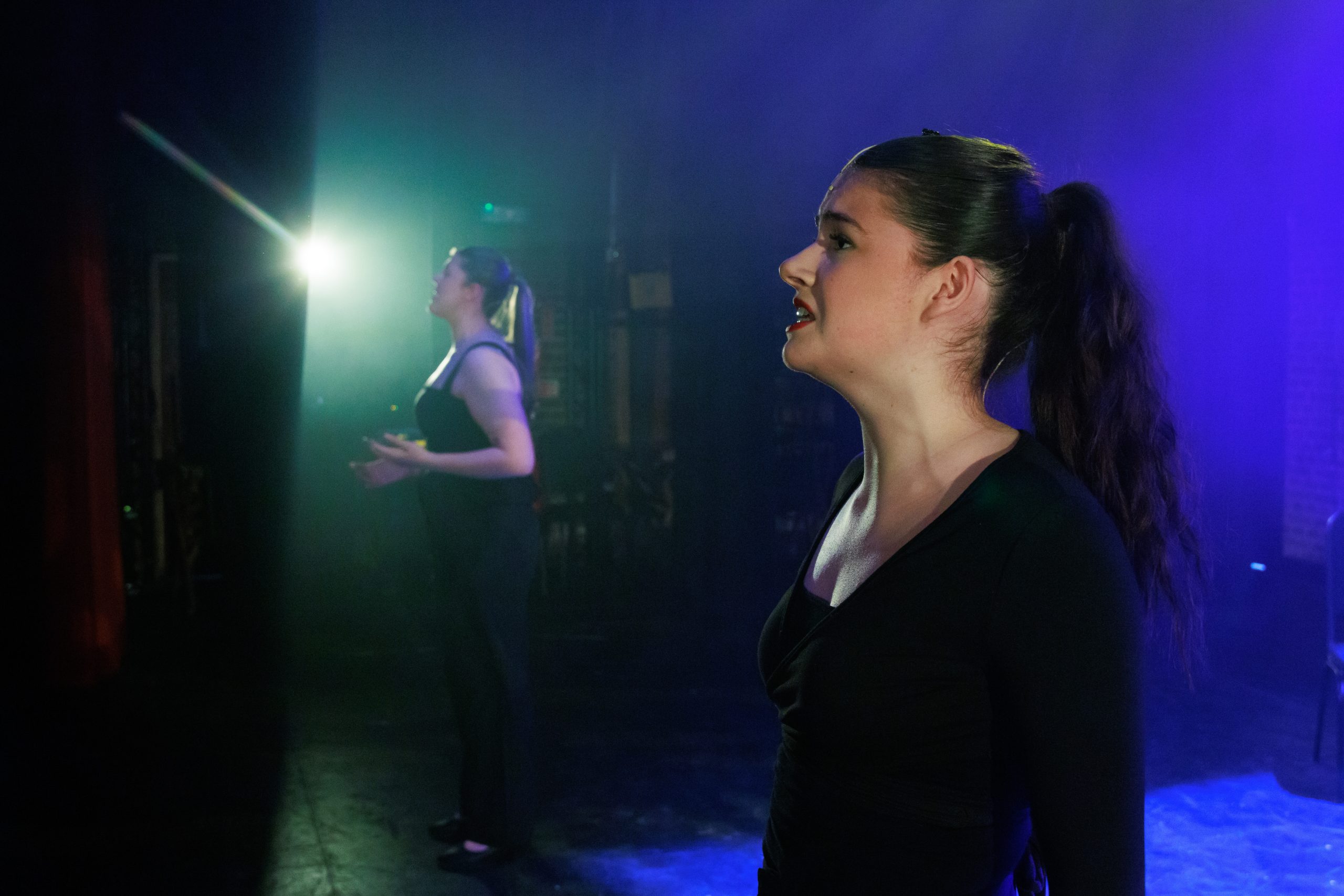 A young woman with a ponytail sings in profile in the foreground, with another performer in green light behind.