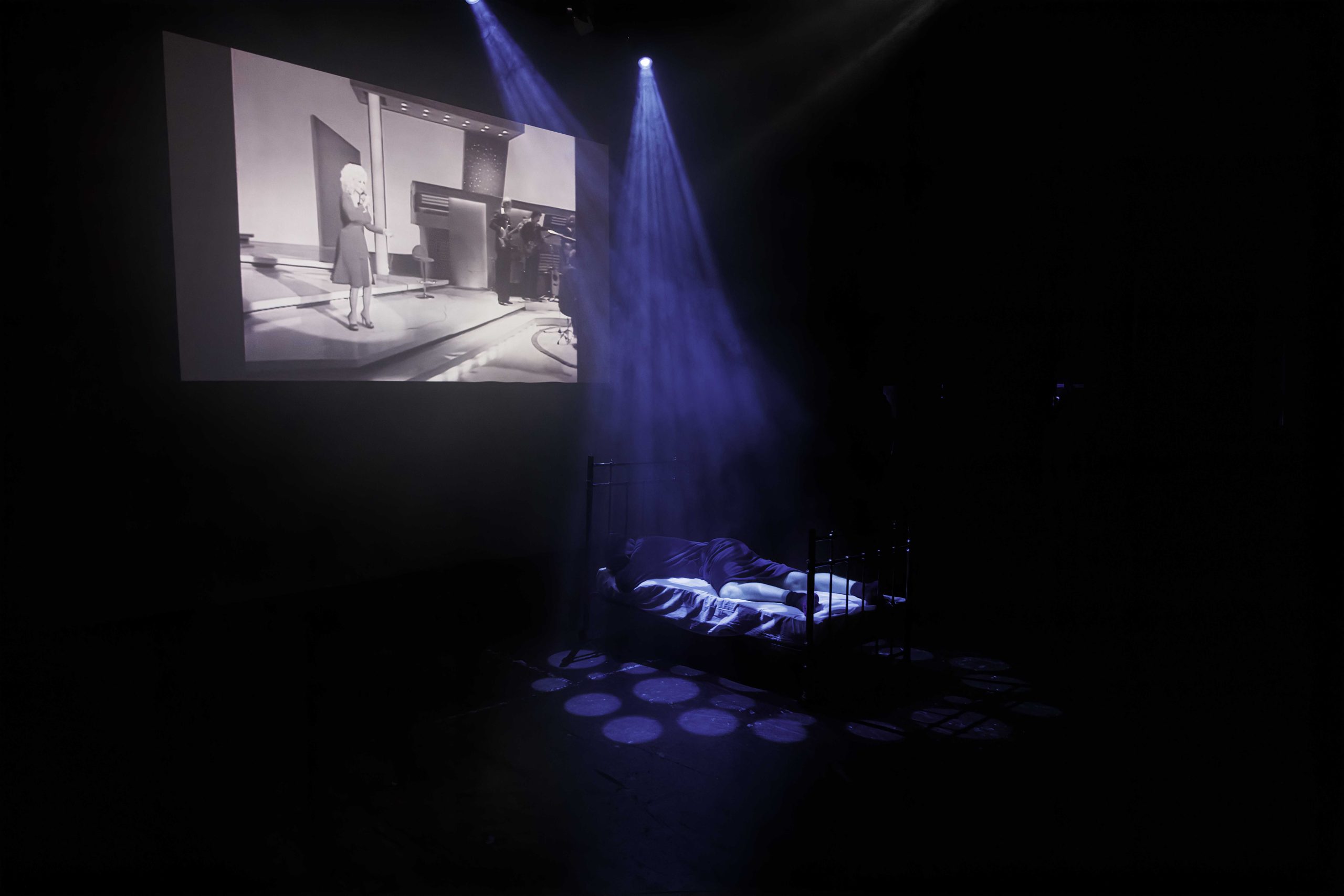 A figure lies on a bed on a dark stage pierced by a beam of light, beneath a black-and-white projection.
