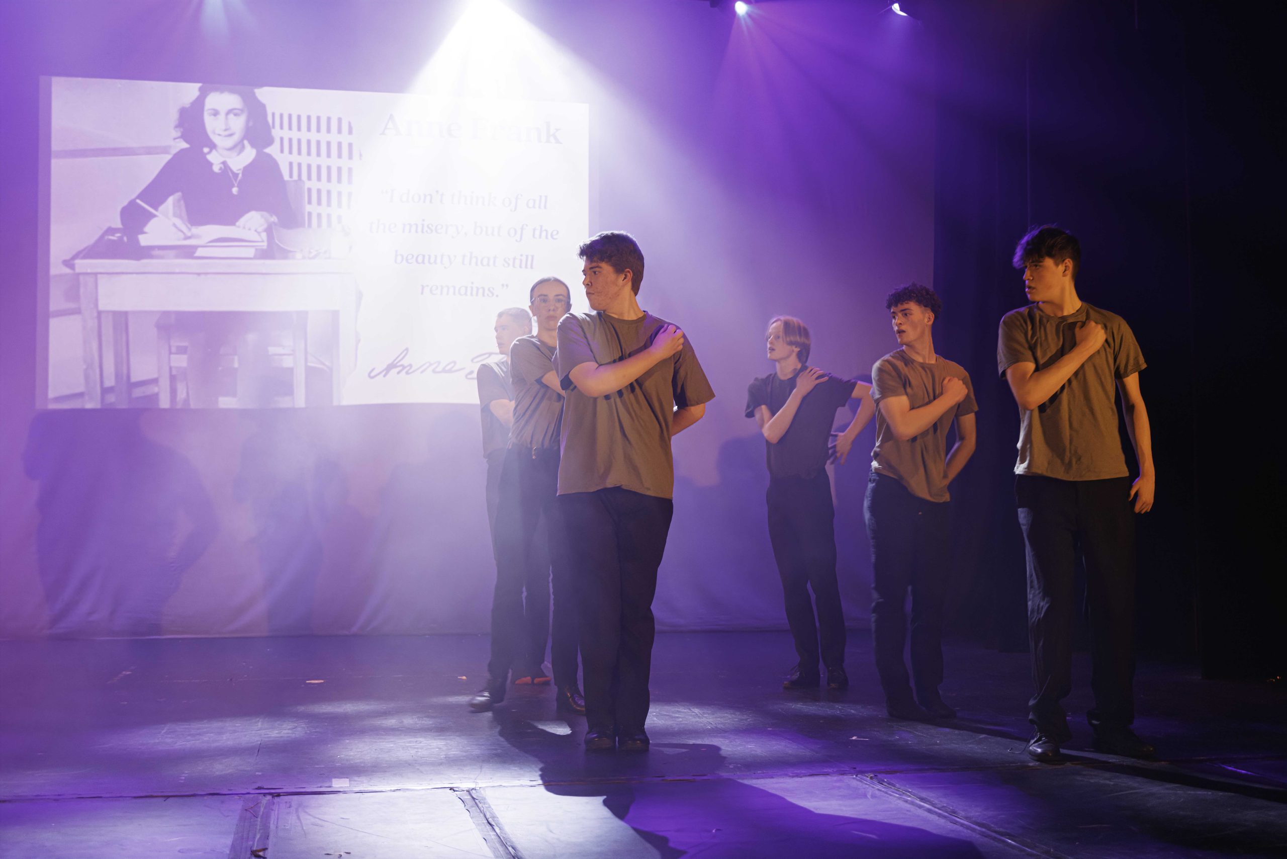 Performers in khaki shirts stand with hands on hearts in front of a projected black-and-white photo of a girl writing at a desk.