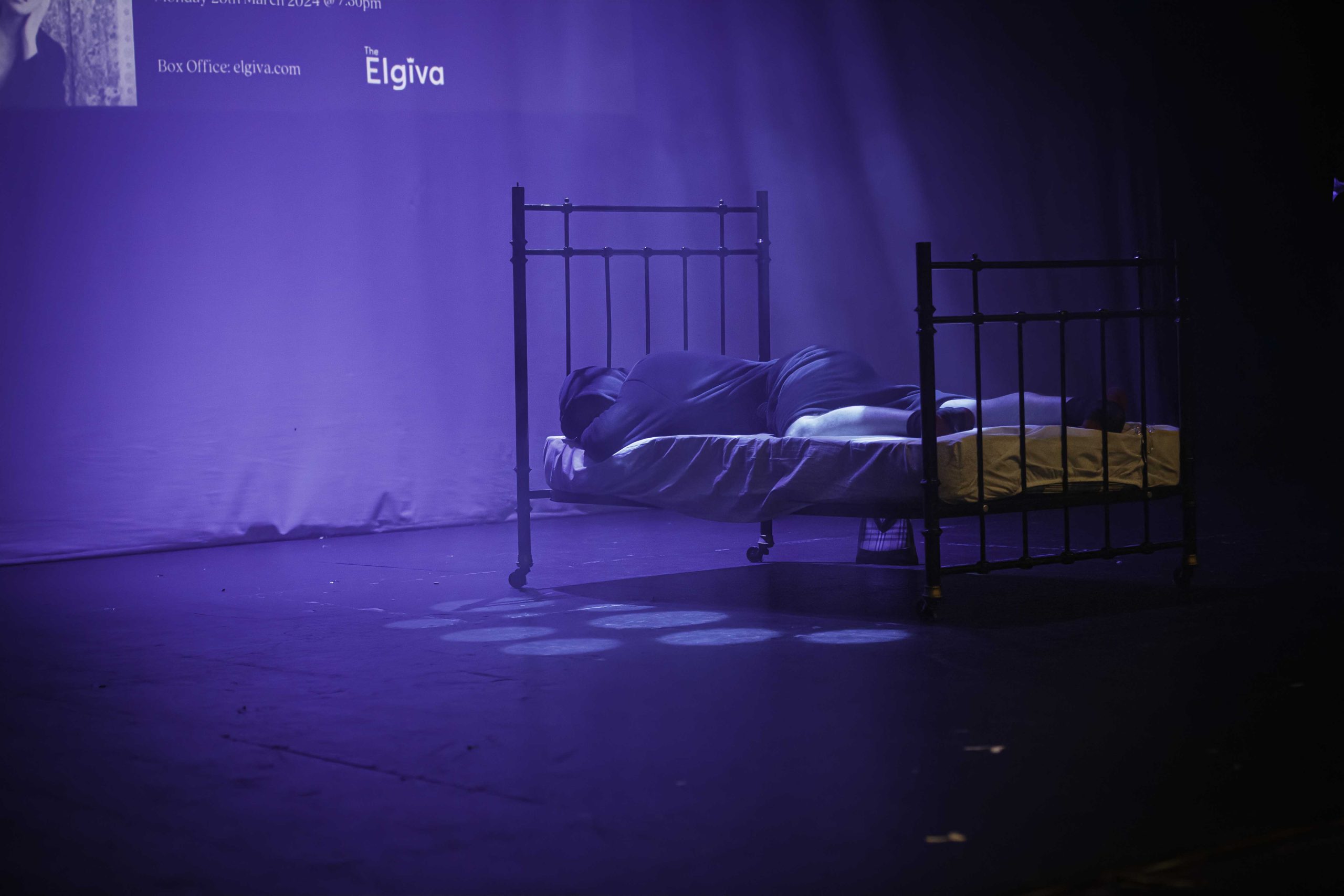 A figure lies under a sheet on an iron bed on a dim purple stage, with a projection screen behind.