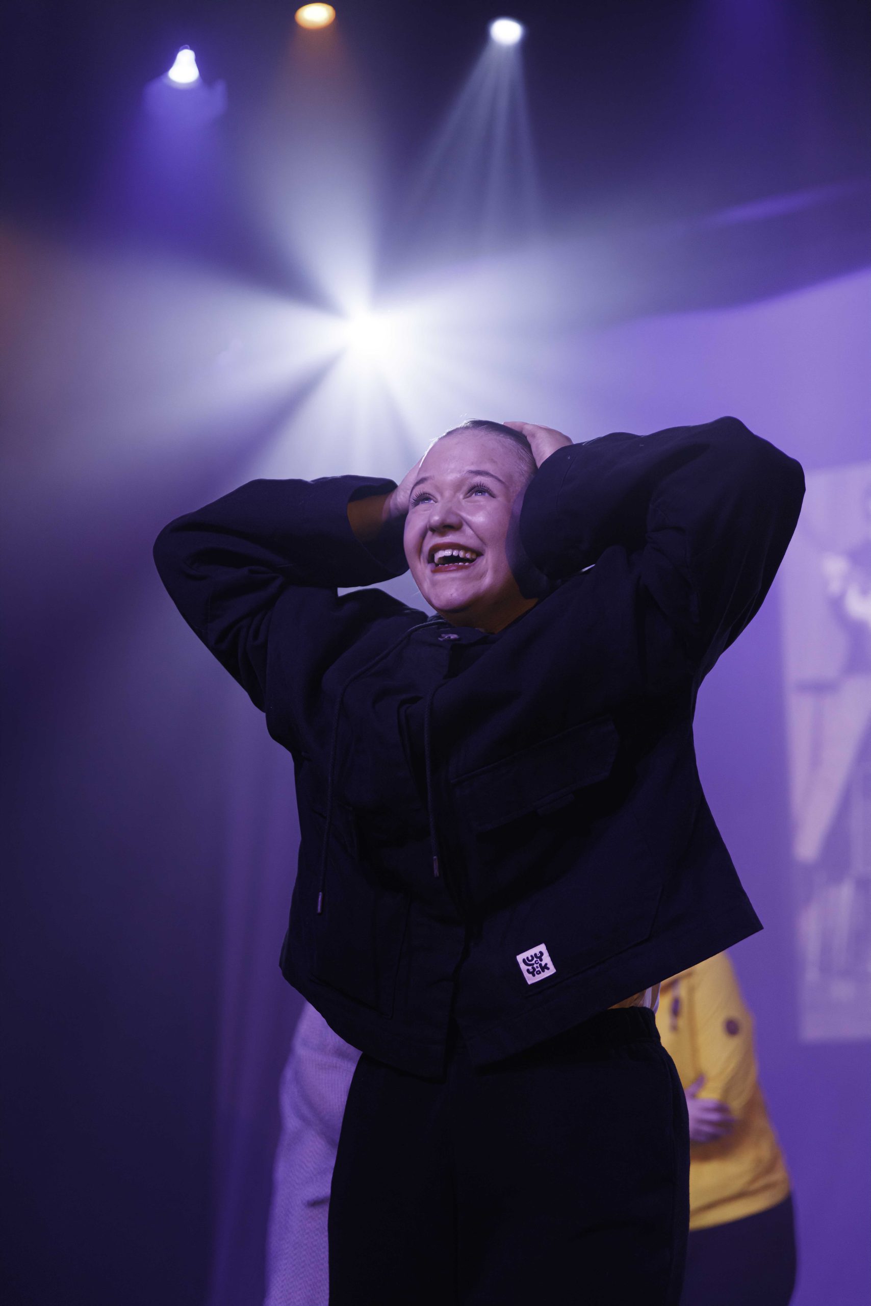 A performer in a dark jacket clasps her head and smiles upward in bright light.