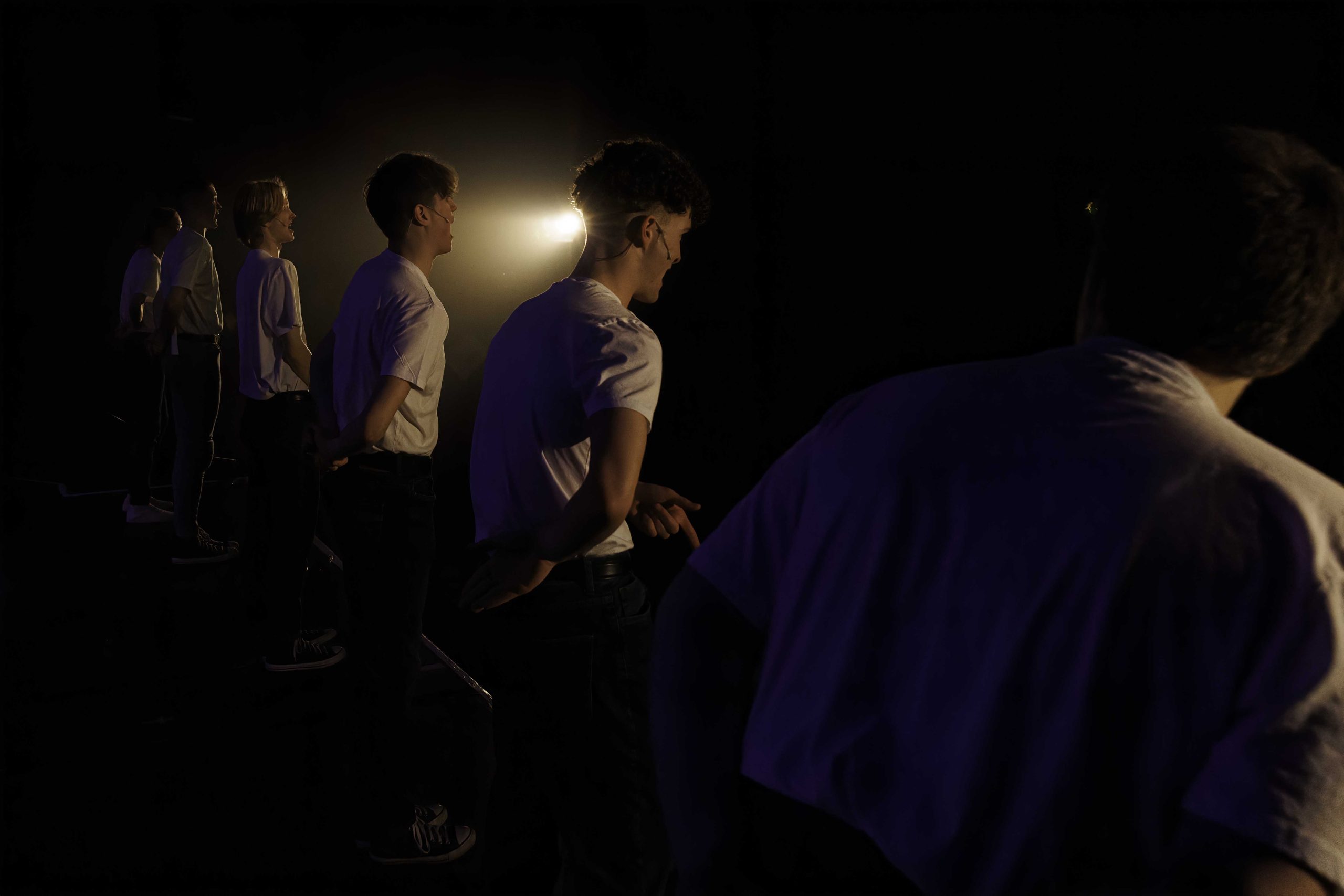 Performers in white T-shirts stand in a dark, hazy space lit by a single side light.