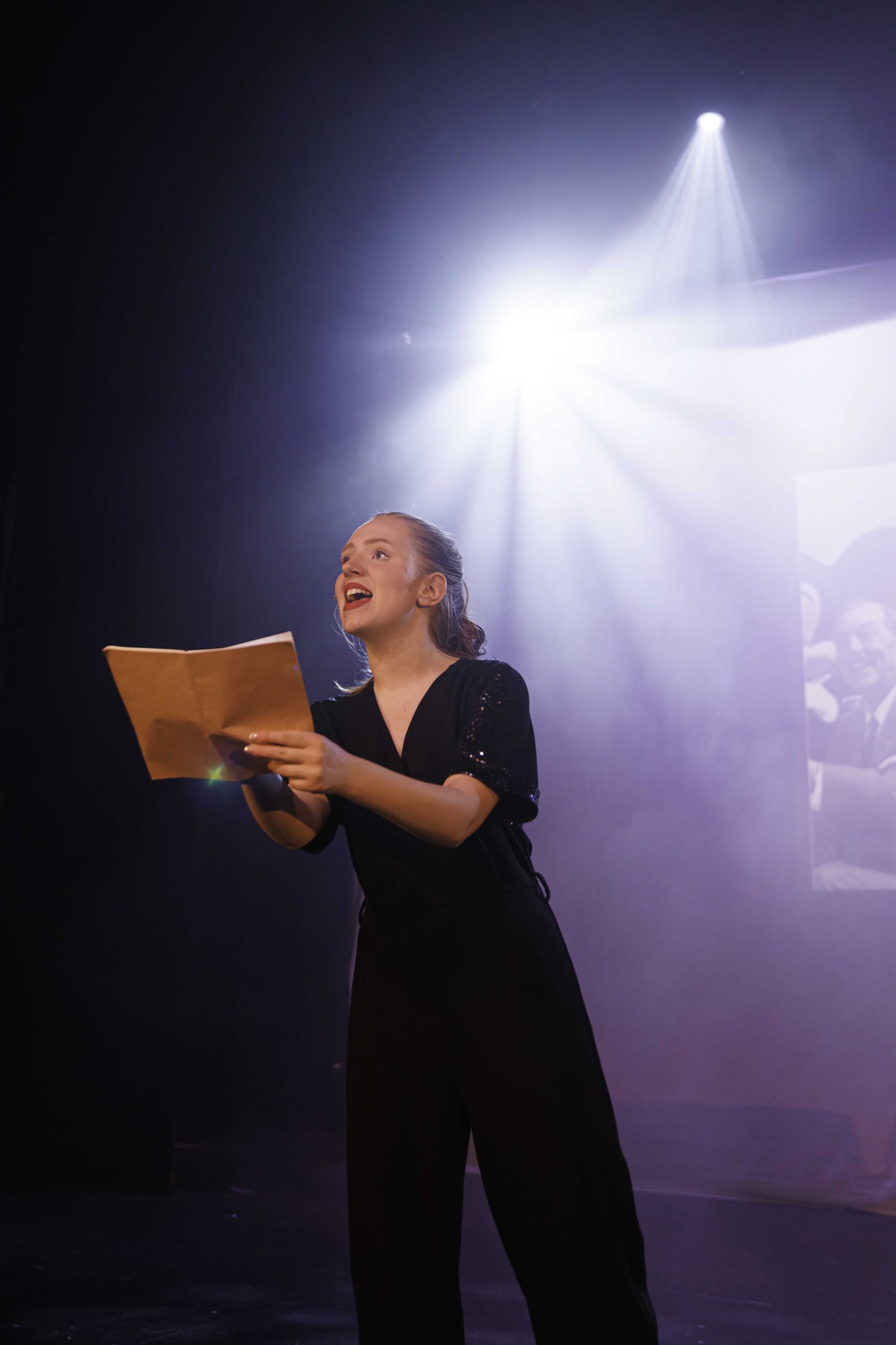 A young woman in black sings while holding an open folder, lit by a bright white spotlight.
