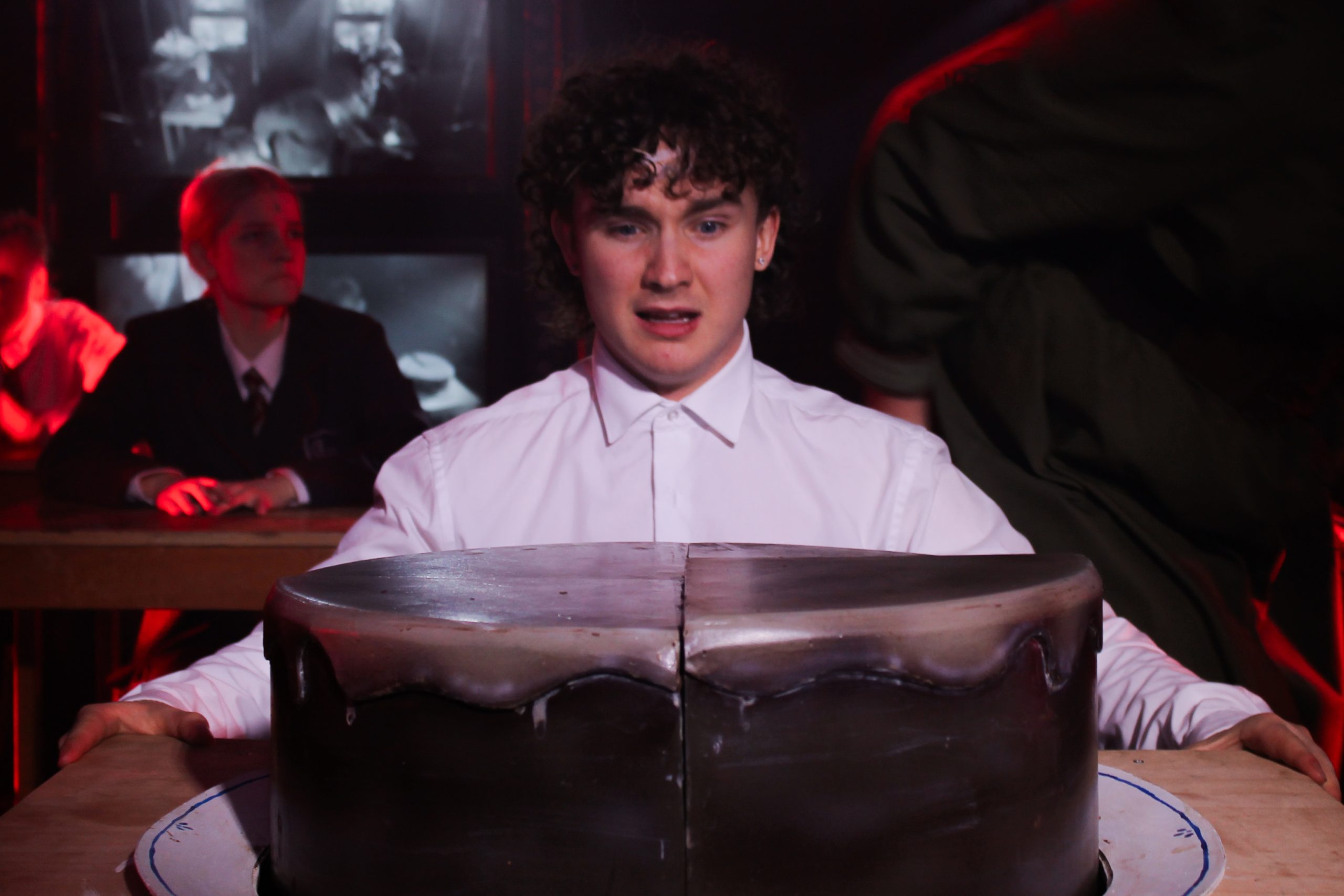 A boy in a white shirt stares at a huge chocolate cake on the desk in front of him.